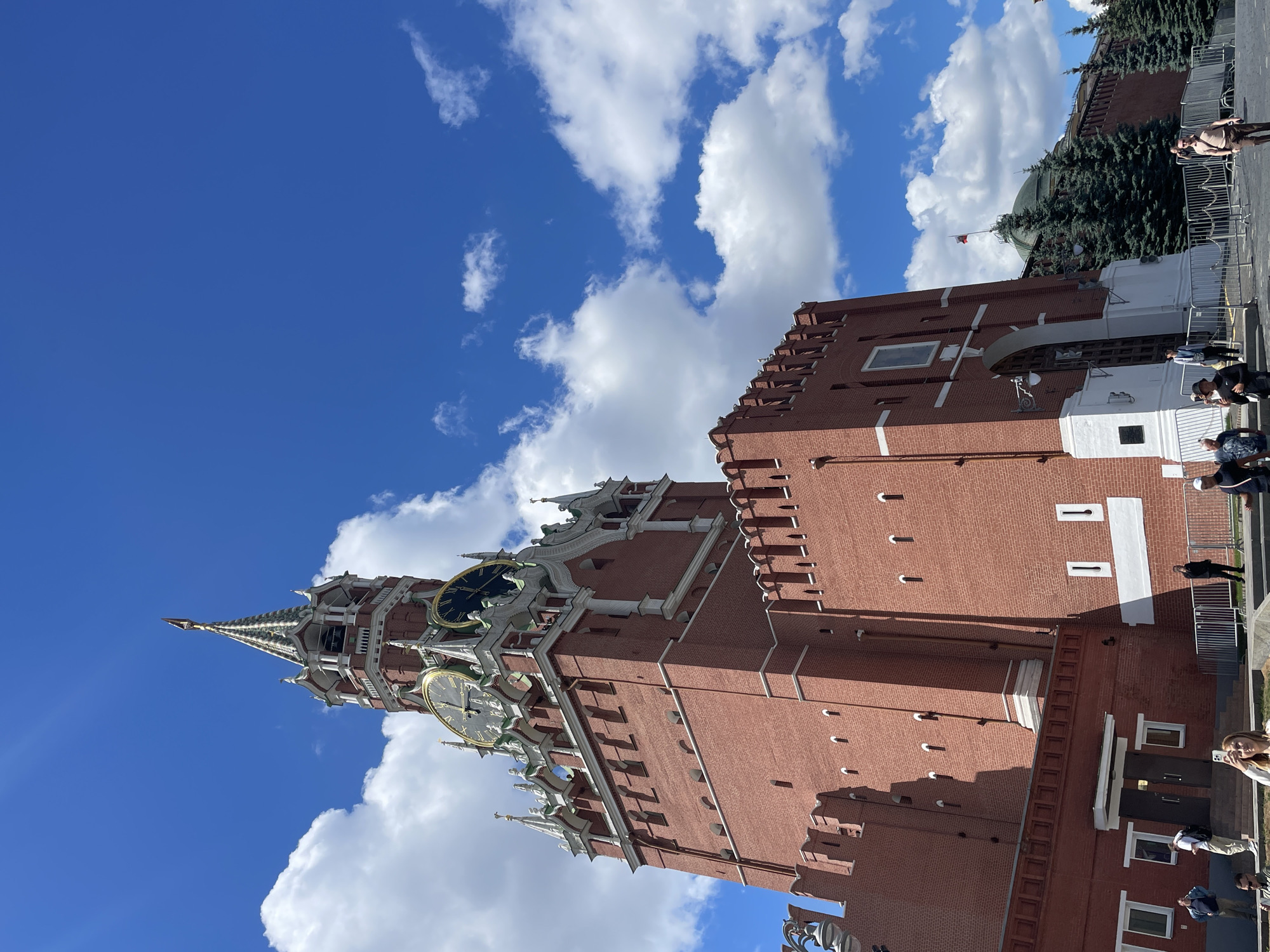 A low-angle, vertical shot shows the Spasskaya Tower of the Moscow Kremlin against a bright blue sky with fluffy white clouds. The tower, constructed of red brick, features a green-domed roof, a prominent clock face, and a spire. Several people are visible in the lower right portion of the image, walking and standing near the tower. Some are captured in the act of taking photos with their phones. The lighting suggests it is daytime, with bright sunlight casting shadows. The overall scene appears to be a public square or tourist area, given the presence of people and the iconic landmark. There is no visible text in the image.