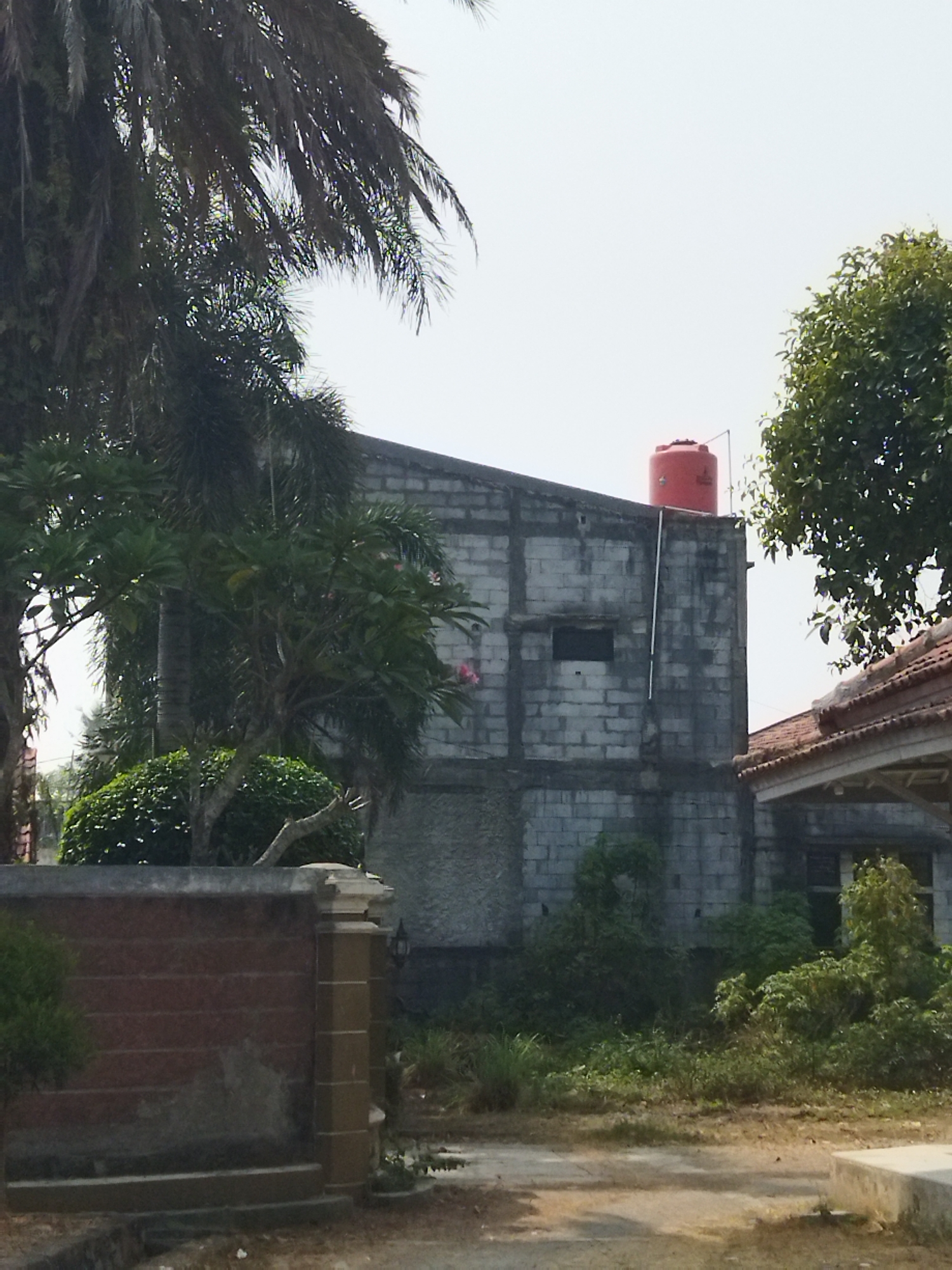 The image depicts a partially constructed building made of concrete blocks in Suka Sari, Indonesia. A large red water tank is visible on the roof of the building. The area around the building is overgrown with vegetation, including palm trees and bushes. In the foreground, there is a brick wall with decorative pillars. The sky is overcast. No people are present in the image.