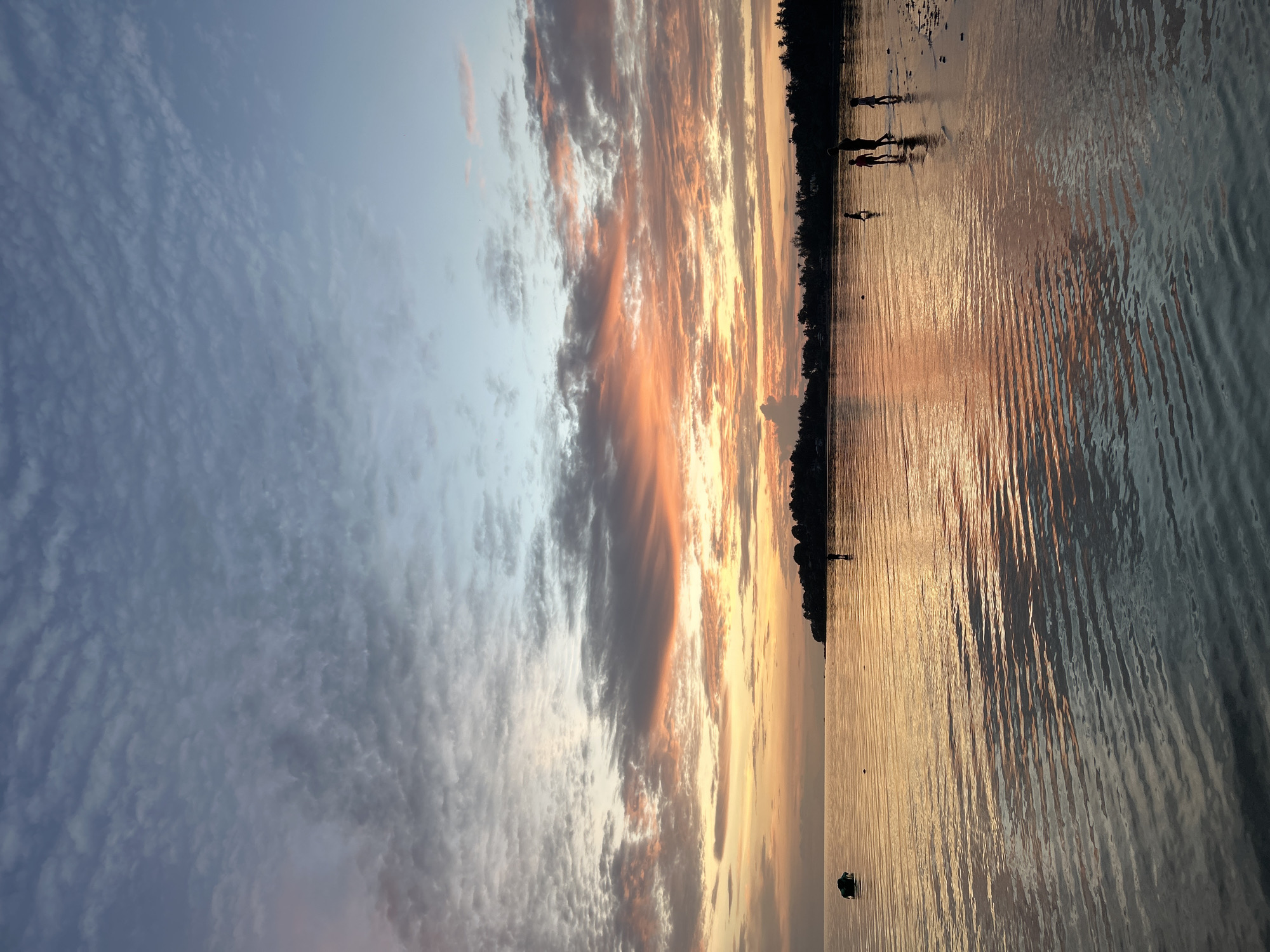 A sunset scene unfolds over the waters of Ko Pha-ngan District, Thailand. The sky is a canvas of soft blues and grays, transitioning to vibrant hues of orange and pink as the sun dips below the horizon. Wispy clouds are scattered across the sky, catching the last rays of light. The calm water reflects the colors of the sky, creating a shimmering surface.
In the foreground, several silhouetted figures are visible standing in the shallow water. They appear to be children and adults, some wading, others walking along the shoreline. A lone boat is anchored in the distance on the left side of the frame. A dark, tree-lined landmass with a subtle slope is visible on the right horizon, indicating the coast. The overall impression is one of a peaceful evening at the beach.