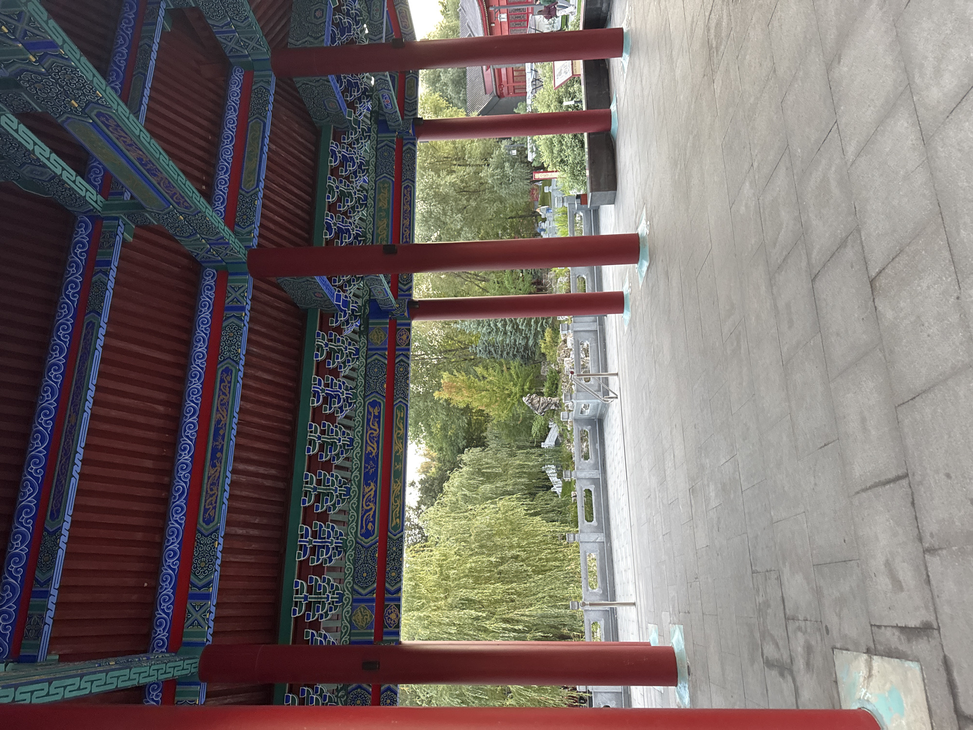 The image shows a covered walkway with ornate Chinese architectural details. Large red pillars support a red wooden roof with intricate blue and green decorative elements. Beyond the walkway, a garden is visible with lush green trees, including a weeping willow, a stone bridge, and a rock formation. The ground in front of the walkway is paved with large gray stone tiles. In the background, a red building with Chinese architectural features can be seen. A few people are scattered throughout the garden. The scene is set in a serene, possibly park-like environment.