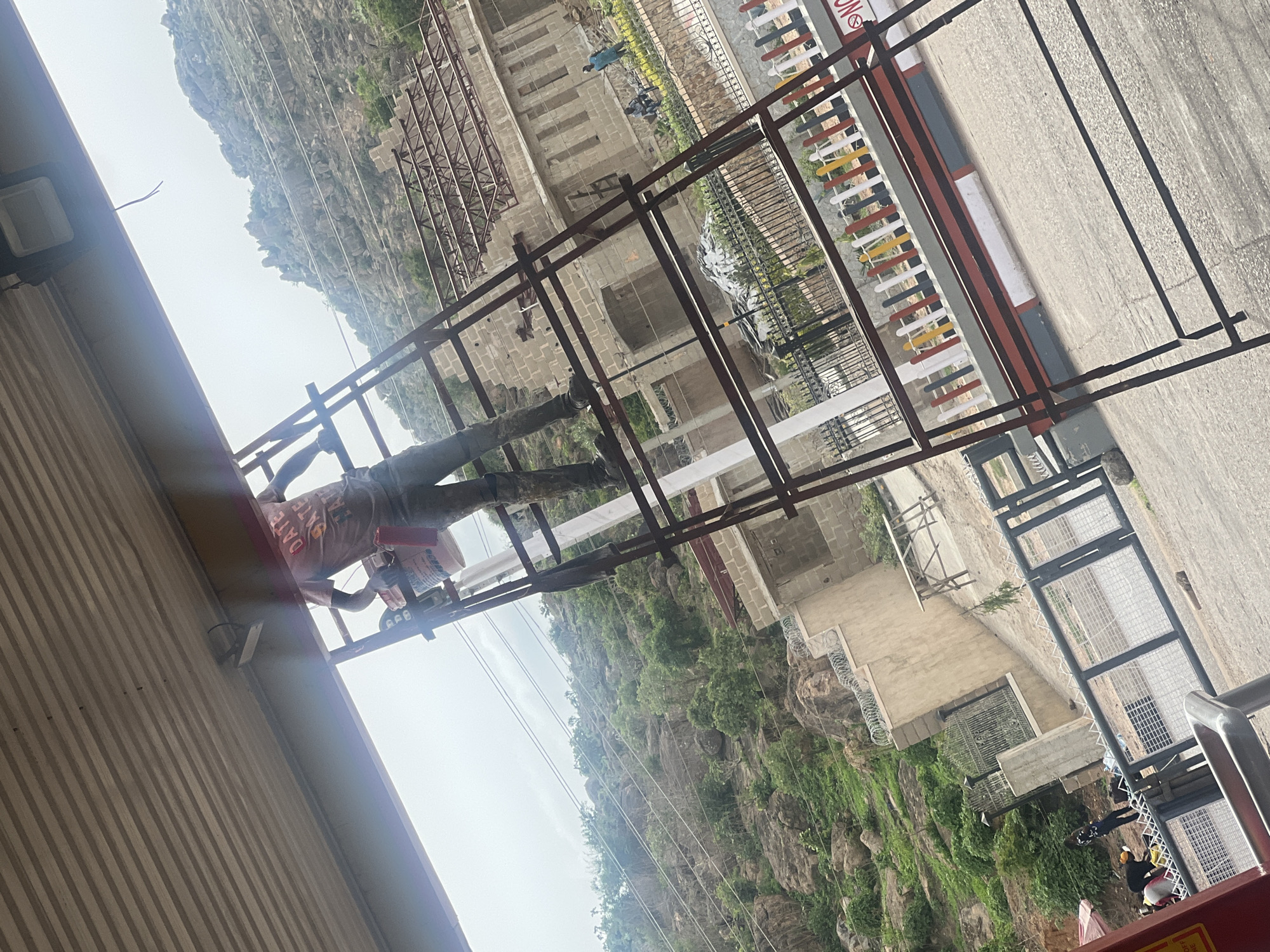 The image depicts a worker painting a metal structure in Gura Top, Nigeria. The worker is standing on scaffolding and appears to be applying paint from a bucket. The background shows a landscape with rock formations and vegetation. A building complex is also partially visible.