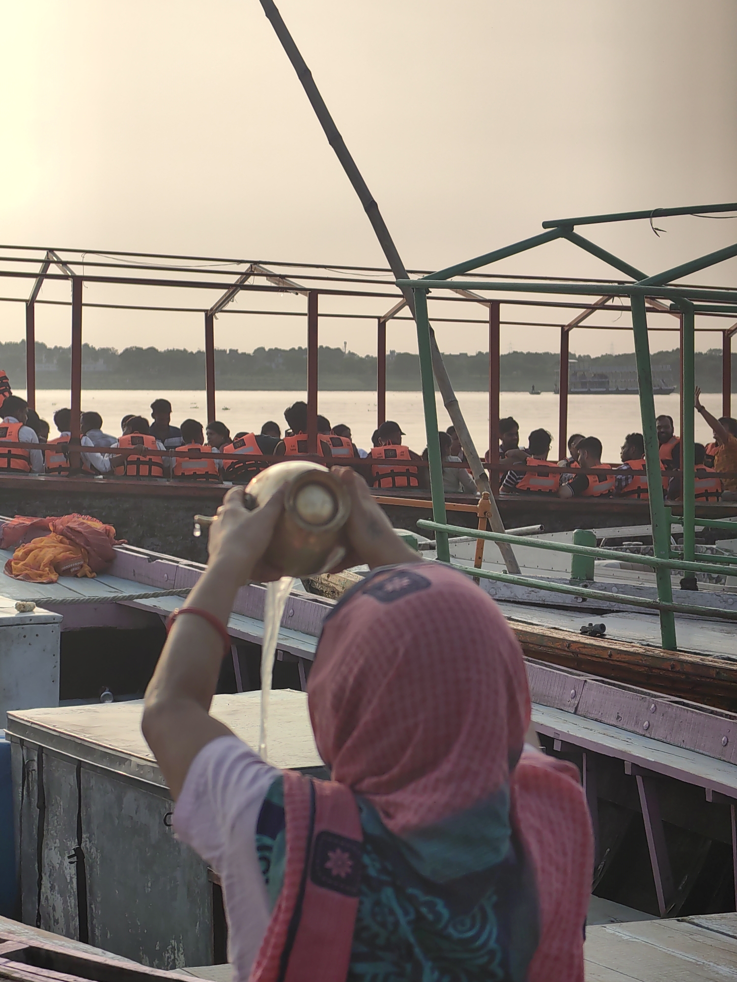 In Varanasi, India, a person is seen from behind, wearing a pink headscarf and a purple and pink patterned shirt, pouring water from a brass pot into the water. The person is standing on a boat. In the background, another boat is filled with people wearing orange life jackets, looking out at the water. The sky is a soft yellow, suggesting either sunrise or sunset. The boats appear to be on a river, likely the Ganges River, a significant site in Varanasi. The scene depicts a moment of reverence or ritual, with the pouring of water being a common practice in religious ceremonies in India. The presence of life jackets suggests a tourist or transport activity on the river.