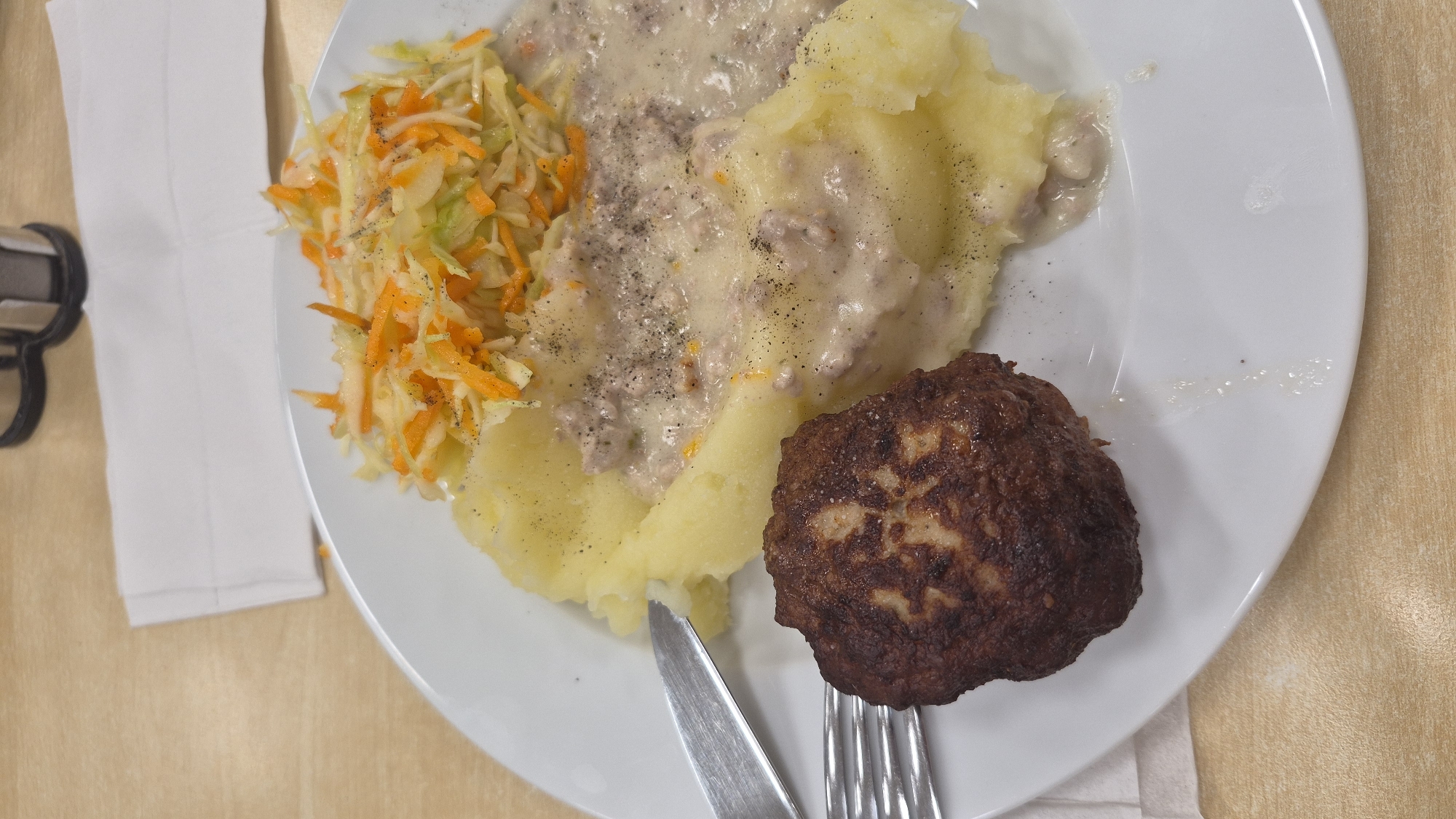 A close-up, overhead shot captures a plate of food on a light brown, wooden table. The plate, made of white ceramic, contains a meal consisting of mashed potatoes, a dark brown meat patty, and a salad. The mashed potatoes form a base for the dish, partially covered by a creamy sauce with visible pieces of meat and black pepper. To the left of the mashed potatoes sits a round, well-browned meat patty with a slightly textured surface. At the top of the plate, a colorful salad composed of shredded cabbage and carrots is piled. A fork rests on the lower left side of the plate, partially under the meat patty, and a knife is positioned on the upper left edge. In the background, a white napkin is folded, and a black object, possibly a salt and pepper shaker set, is visible. The image provides no information about the specific location in Preiļi, Latvia, beyond it being a dining setting.