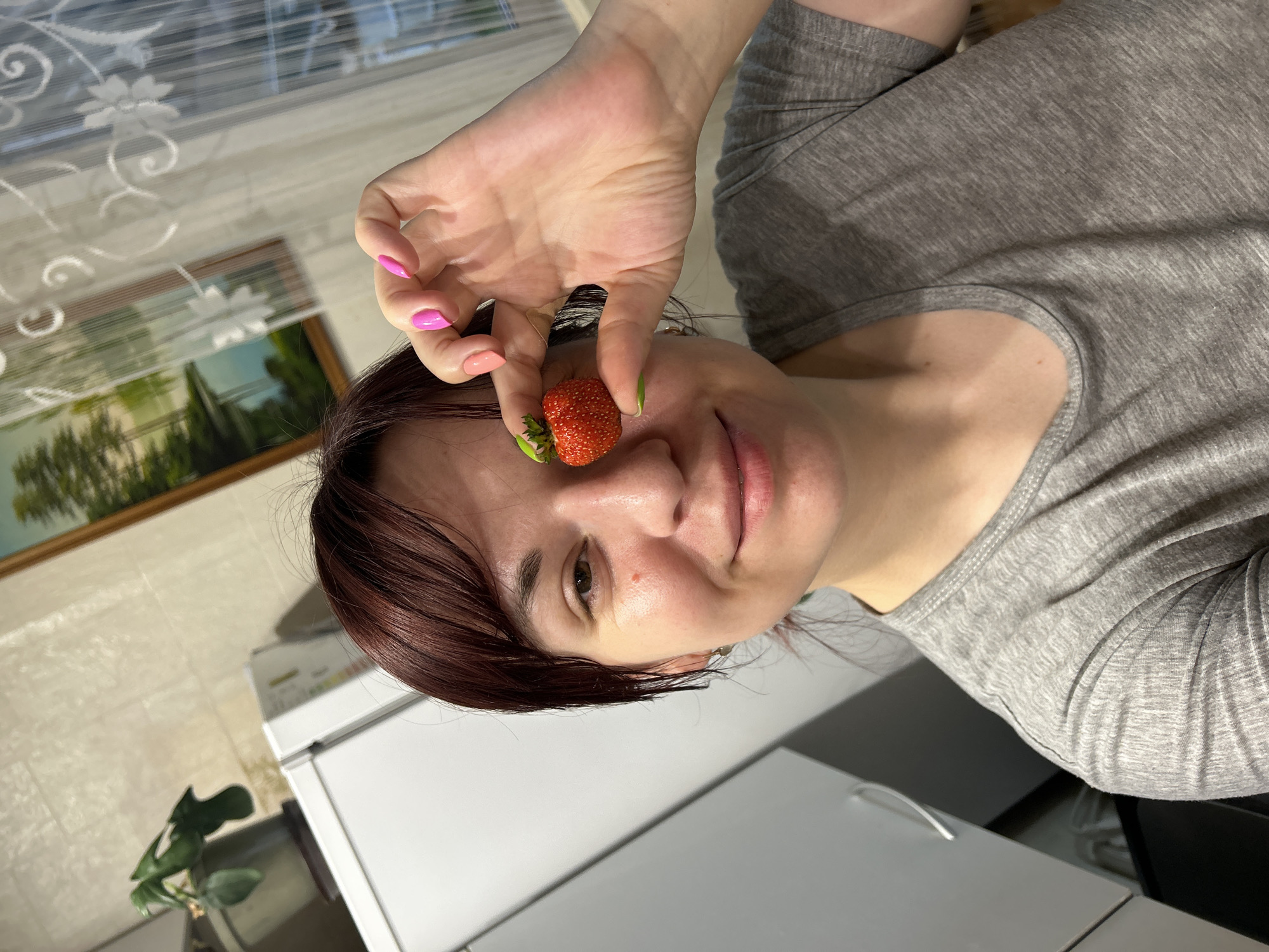 The media file shows a close-up, rotated view of a woman holding a strawberry up to her face, near her eye. The woman has dark hair and is wearing a grey t-shirt. Her fingernails are painted in shades of pink and green. She is smiling slightly. The background appears to be a domestic setting, possibly a kitchen, with white appliances visible. There is also a framed picture on the wall and a houseplant. The lighting suggests it is daytime. The overall mood is playful and casual. No visible text is present.