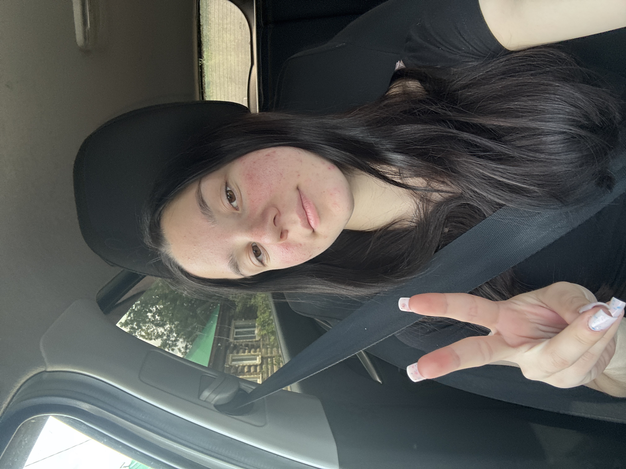 The media file is a vertically oriented, close-up selfie taken by a young woman in a car. She is wearing a black t-shirt and a black hat. Her long, dark hair is visible around her face and shoulders. She has light brown eyes and her face shows some redness and blemishes. Her lips are slightly parted, and she is looking towards the camera with a neutral expression. Her right hand is raised, and she is making a peace sign with her index and middle fingers extended. Her nails are long and painted with a light-colored polish that has a speckled or glittery design.
The background suggests the car is in motion, with trees and a building visible through the passenger window. The lighting indicates it is daytime, likely with overcast or diffused sunlight. The scene is set within the interior of a vehicle, indicated by the car seat, seatbelt, and interior paneling. The presence of the seatbelt suggests the woman is either the driver or a passenger.
Notable details include the woman's facial features and the detail of her manicured nails. The background provides a glimpse of an outdoor environment, possibly a suburban or rural setting given the trees and building. There is no visible text in the media file.