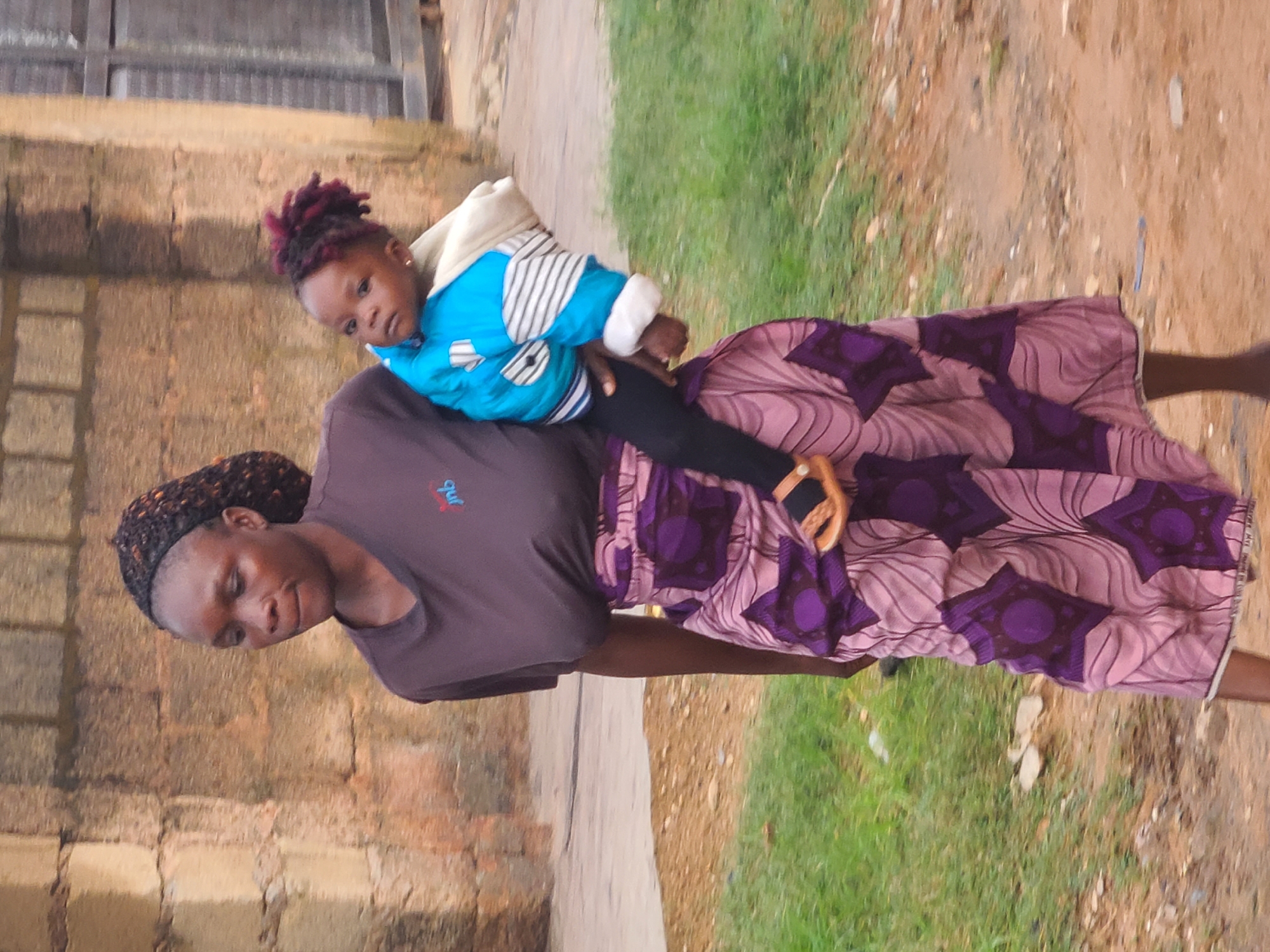 In the city of Jos, Nigeria, a woman with braided hair is walking outdoors. She is carrying a young child on her left hip. The child is wearing a blue jacket with white stripes and a light-colored hood. The woman is wearing a dark-colored t-shirt with a small logo on the chest and a long, patterned skirt in shades of pink and purple. She has a sandal on her right foot, and the child's left foot is also visible with a sandal. They are walking on a dirt path with green grass to the left and a textured brick wall in the background. The overall scene suggests an everyday moment in an outdoor urban environment.