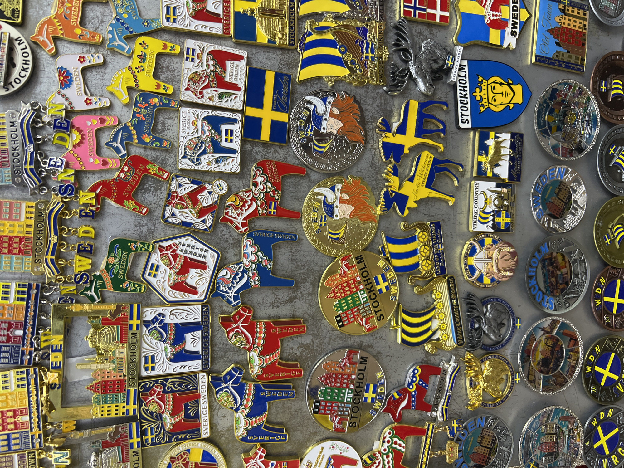 The image displays a collection of Swedish-themed souvenirs, primarily magnets, featuring iconic imagery of Stockholm and Swedish culture. The subjects are mostly decorative items, including Dala horses in various colors, Viking figures, moose, and representations of the Stockholm skyline with its distinctive colorful buildings and churches. Several items also bear the Swedish flag. The souvenirs are arranged in a dense, overlapping pattern against a textured, grey background, likely a refrigerator or magnetic board. The overall impression is one of a curated display of tourist memorabilia.