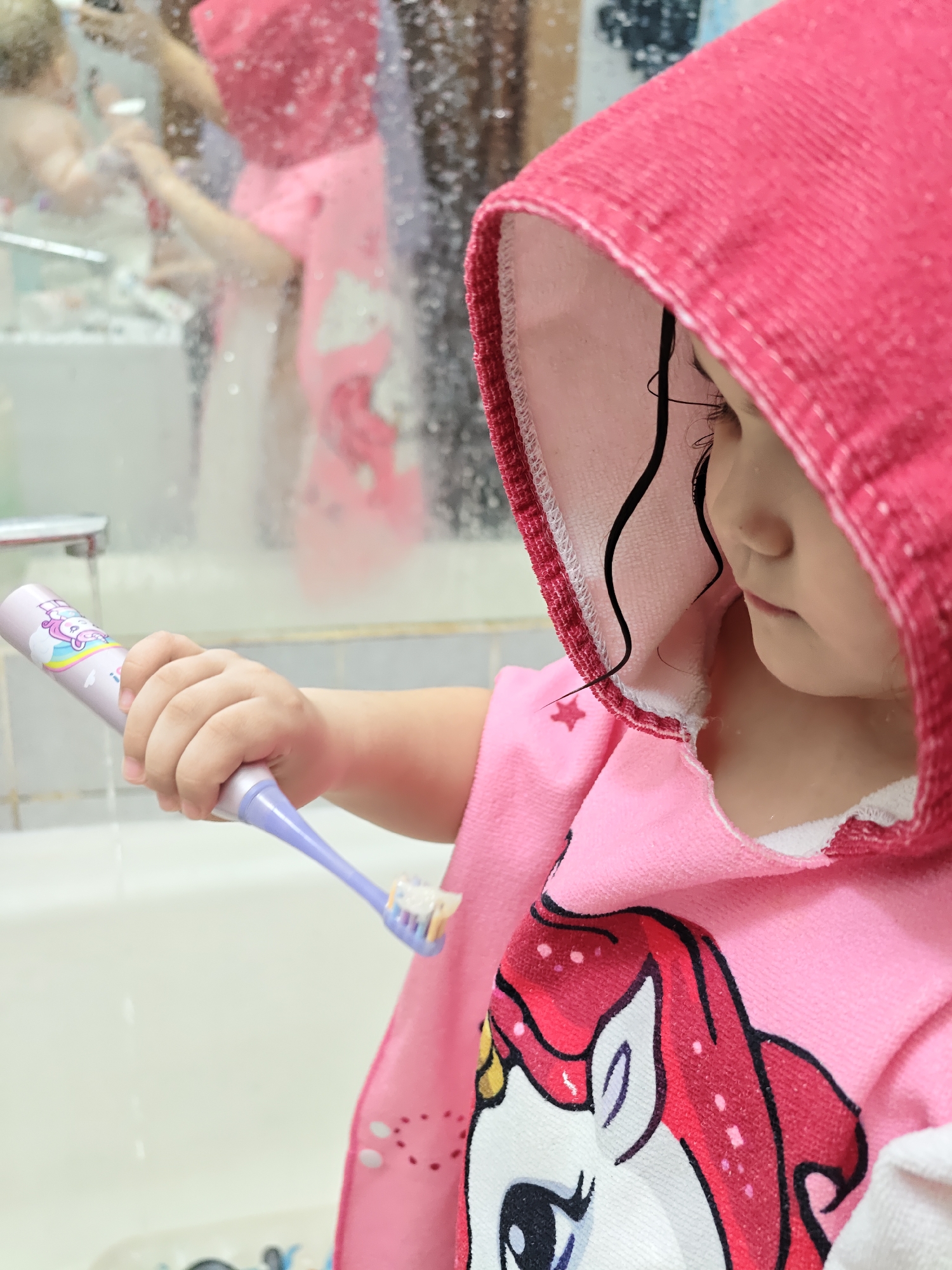 A young child in a pink hooded towel, adorned with a unicorn design, is holding a purple toothbrush under running water in a bathroom. The child is fair-skinned with dark hair partially visible under the hood. In the background, a person reflected in the bathroom mirror appears to be wearing a similar pink towel. The bathroom features a white sink and faucet. The image does not reveal any location information about Chaikovsky, Russia.