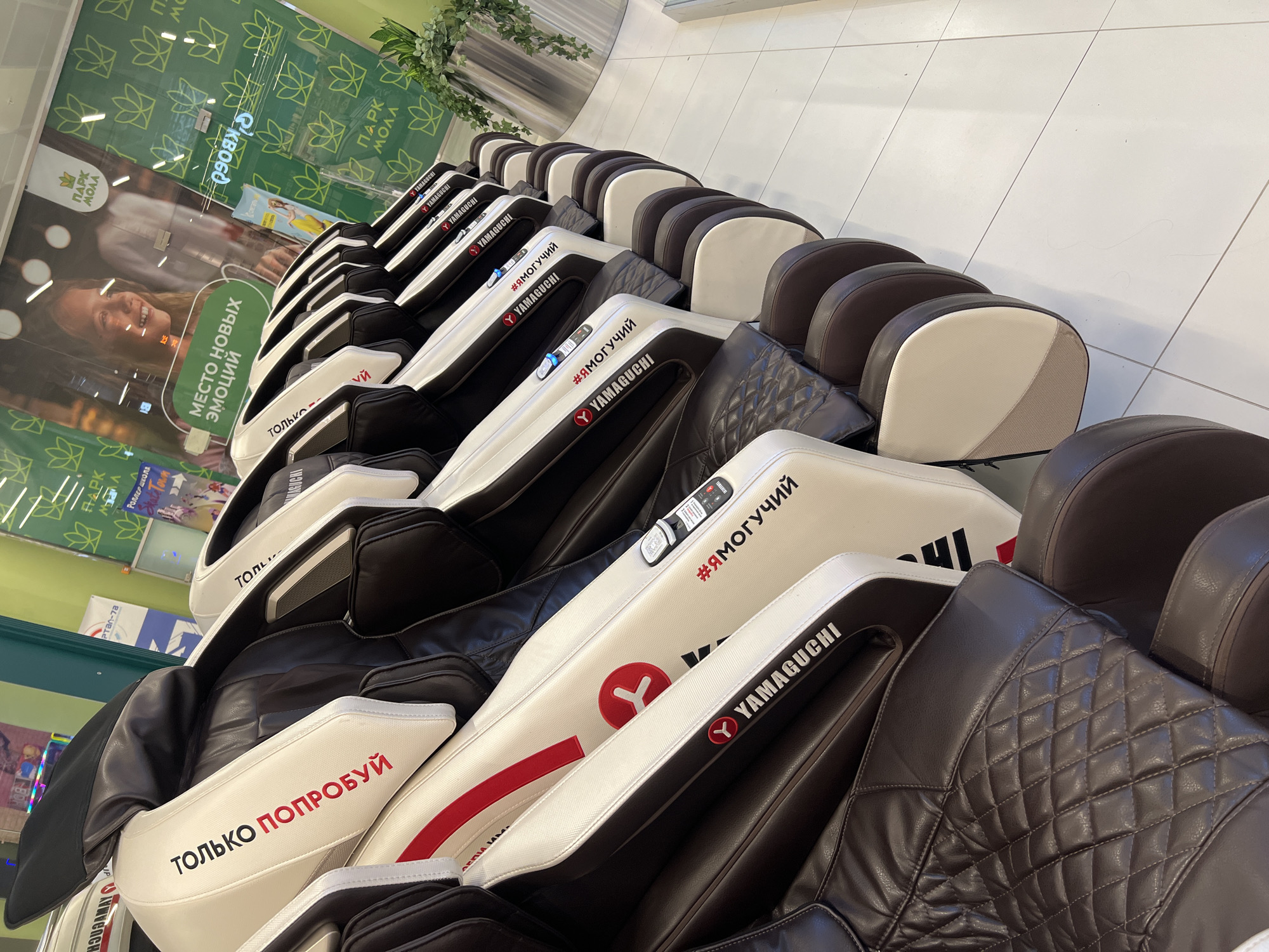 The image shows a row of Yamaguchi massage chairs, likely in a retail store. The chairs are predominantly white and dark brown, with some red accents. Text in Russian is visible on the side of the chairs, including "ТОЛЬКО ПОПРОБУЙ" (Just Try) and "#ЯМОГУЧИЙ" (#IAmPowerful). The chairs are lined up in a retail space with a tiled floor and a large window or wall display in the background. The display features a young girl smiling and text that appears to be an advertisement. The overall environment suggests a commercial setting. There are no people clearly visible in the image. The location is identified as Irinovka, Russia, though this specific detail is not visually verifiable from the image itself but is provided as context.