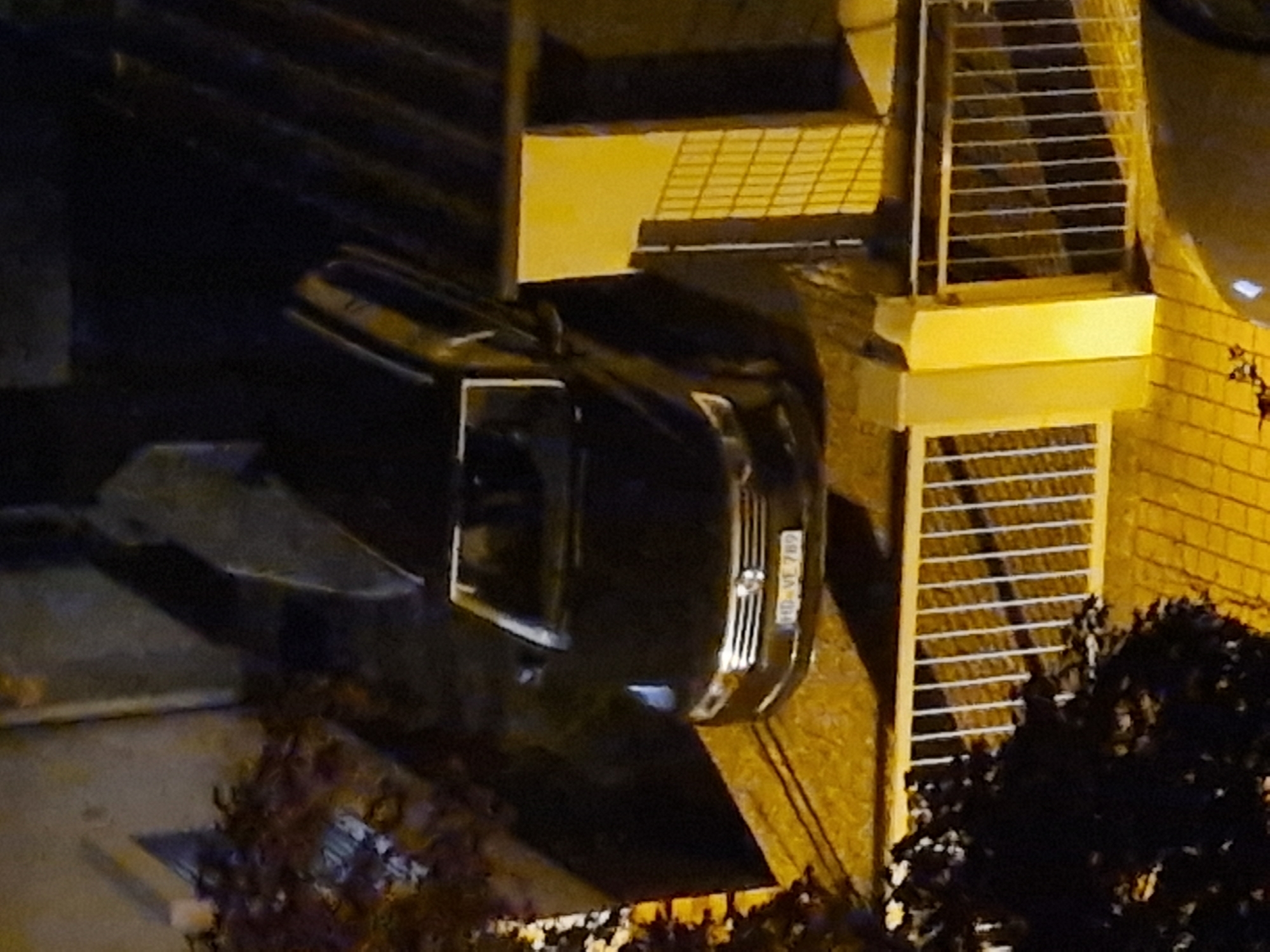 The image shows a black station wagon parked on an elevated area with a yellow patterned surface. The car's license plate is visible and reads "HD-VE 789". A white metal fence runs along the bottom of the image, separating the car's parking area from a lower section of pavement. There is a shadowy figure in the background, partially obscured by darkness and what appears to be a roof structure. The overall lighting suggests it is nighttime, with artificial light sources casting yellow hues. The location is identified as Leimen, Germany.