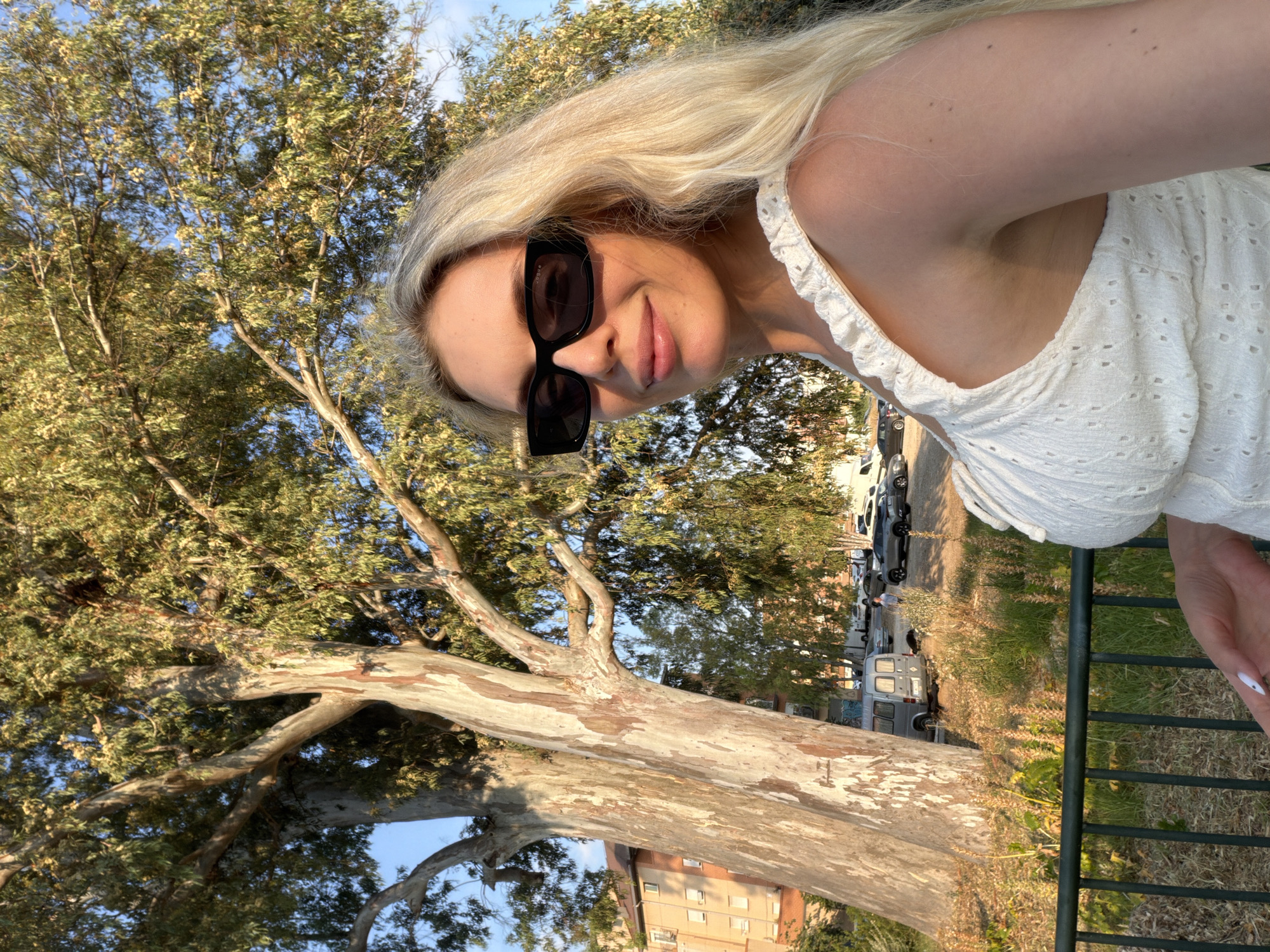 A blonde woman wearing sunglasses and a white eyelet top is posing for a selfie outdoors. She has long, blonde hair and is smiling. Behind her, a large tree with textured bark is prominent, with a building and several parked vehicles visible in the background. The scene appears to be in a park-like setting, possibly in Benimàmet-beniferri, Spain, indicated by the presence of Mediterranean vegetation and architecture. The lighting suggests it is late afternoon or early evening.