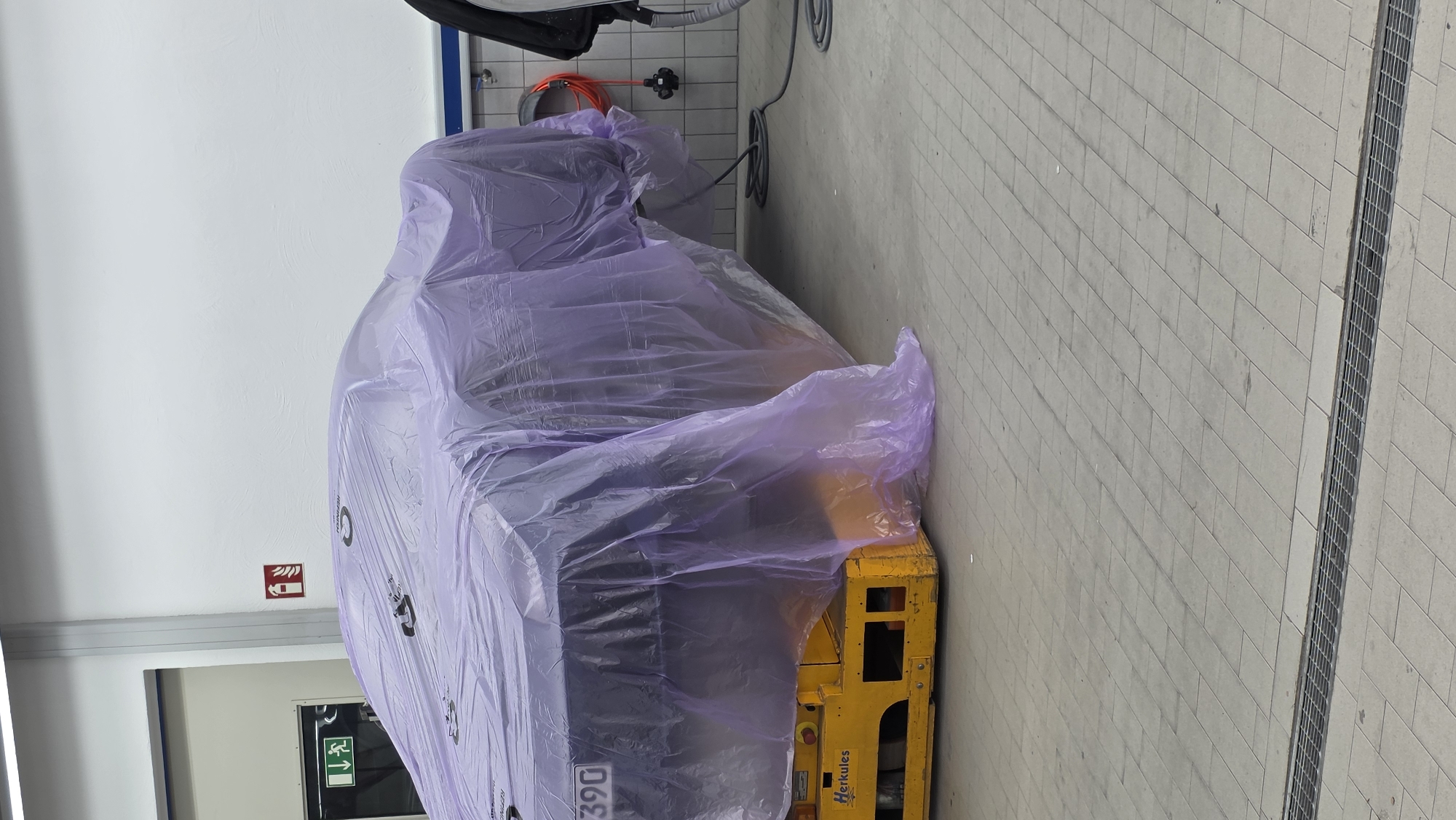 A vehicle, draped in a translucent purple plastic sheet, is positioned on a yellow industrial trolley. The trolley has the word "Herkules" printed on its side. The vehicle appears to be in a workshop or garage setting, with a tiled floor and a white wall in the background. A fire extinguisher symbol and an exit sign are visible on the wall. Electrical cords and a vacuum cleaner are also present in the scene. The overall lighting suggests an indoor environment.