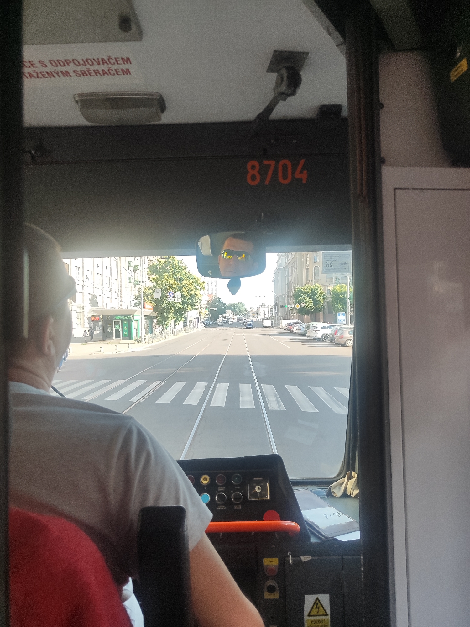 The image is taken from the perspective of a tram driver, looking forward through the windshield and a rearview mirror. The tram appears to be in motion, proceeding down a street with tram tracks. In the rearview mirror, the driver's face is visible, wearing sunglasses. The street outside features crosswalks, parked cars, buildings, and trees. The interior of the tram shows the driver's console with various buttons and controls, as well as a red steering wheel. There is text in a foreign language on the tram's ceiling and on a sign in the distance. The overall scene depicts a sunny day in an urban environment.