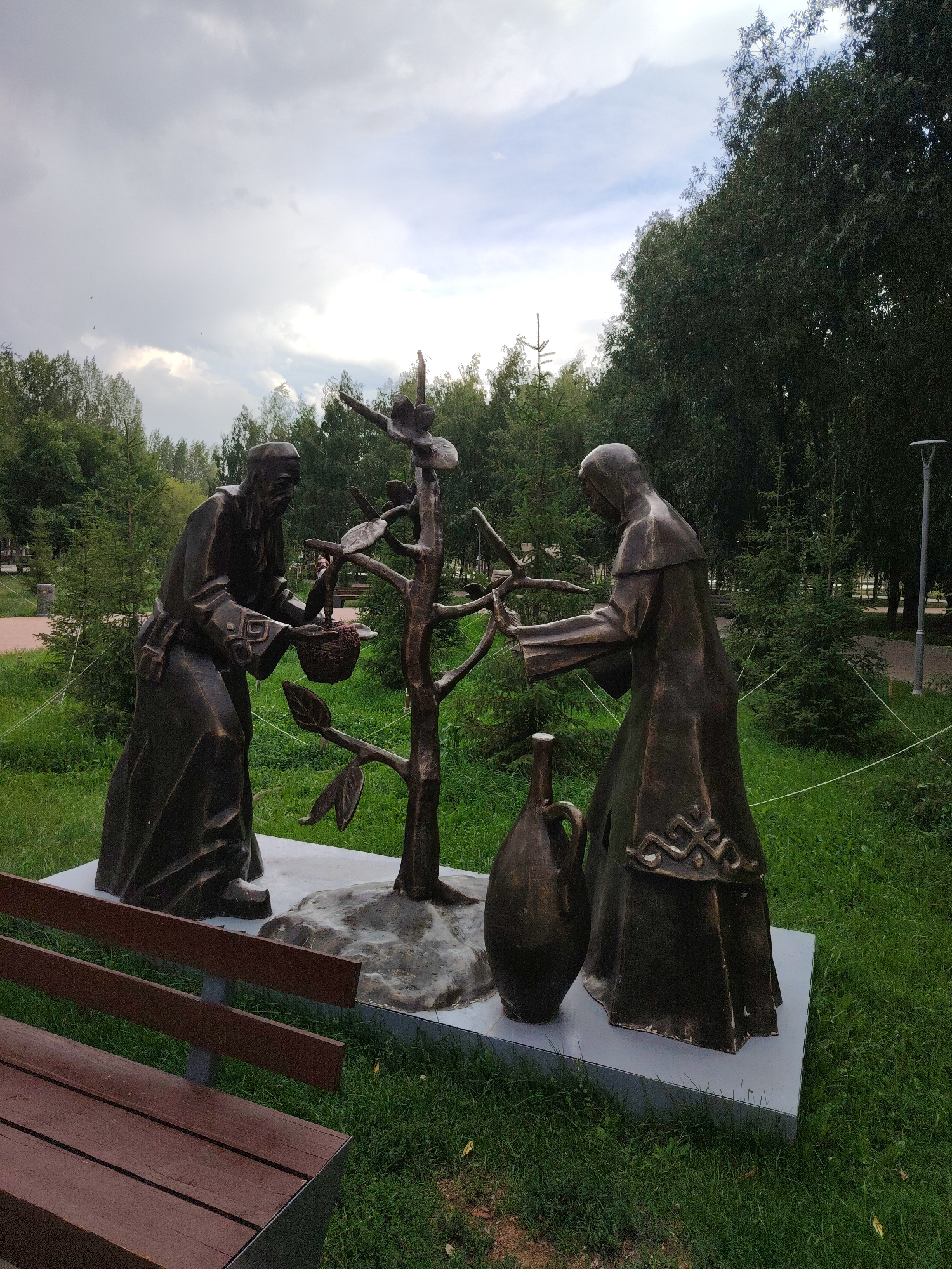 The image depicts a bronze sculpture in a park setting. Two figures, appearing to be historical or folkloric characters, are shown interacting with a stylized tree. The figure on the left, wearing a hooded robe, holds a basket containing what appears to be fruit or berries. The figure on the right, also robed and hooded, reaches towards the tree. Beside the base of the sculpture is a large ceramic jug. The sculpture is situated on a concrete platform, surrounded by green grass and trees, under a cloudy sky. A wooden park bench is visible in the foreground. This sculpture is located in the city of Astana, Kazakhstan, likely within a park or public space, possibly commemorating a historical or cultural event related to agriculture or sustenance.