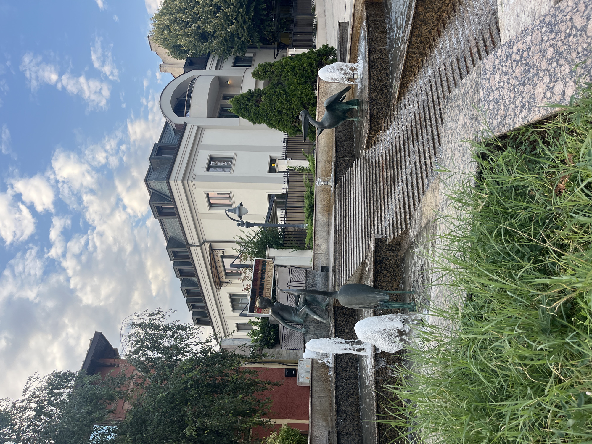 The image depicts a contemporary fountain in the city of Burgas, Bulgaria, located in front of a multi-story building. The fountain features tiered levels with cascading water and granite detailing. Two bronze sculptures are prominently displayed within the fountain: one of a boy sitting and reaching upwards, and another of a bird, possibly a heron or crane. Water jets are active, creating a dynamic visual effect. Lush green grass borders the foreground, and the background shows a modern architectural facade with a mansard roof and dormer windows, partially obscured by trees. The sky above is partly cloudy with a clear blue sky. A street lamp and a fence are also visible in the mid-ground. The overall scene suggests a public urban space with artistic elements.