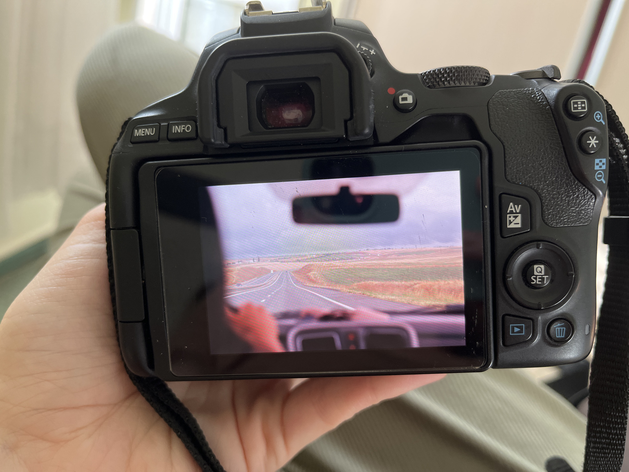 An extreme close-up, overhead shot shows a black DSLR camera being held by a person's hand. The camera screen displays a view from inside a car, looking out through the windshield. The road ahead curves to the left, with fields of green and brown on either side. The interior of the car is visible, including the dashboard and a rearview mirror. The camera's buttons and dials are also visible, including the "MENU," "INFO," "Av," "Q SET," and a playback button. The background is blurred, but appears to be an indoor setting with light-colored curtains and a muted green surface, possibly a couch or chair. The image does not contain any information that would allow for identification of the location as Komsomolskoye, Ukraine.