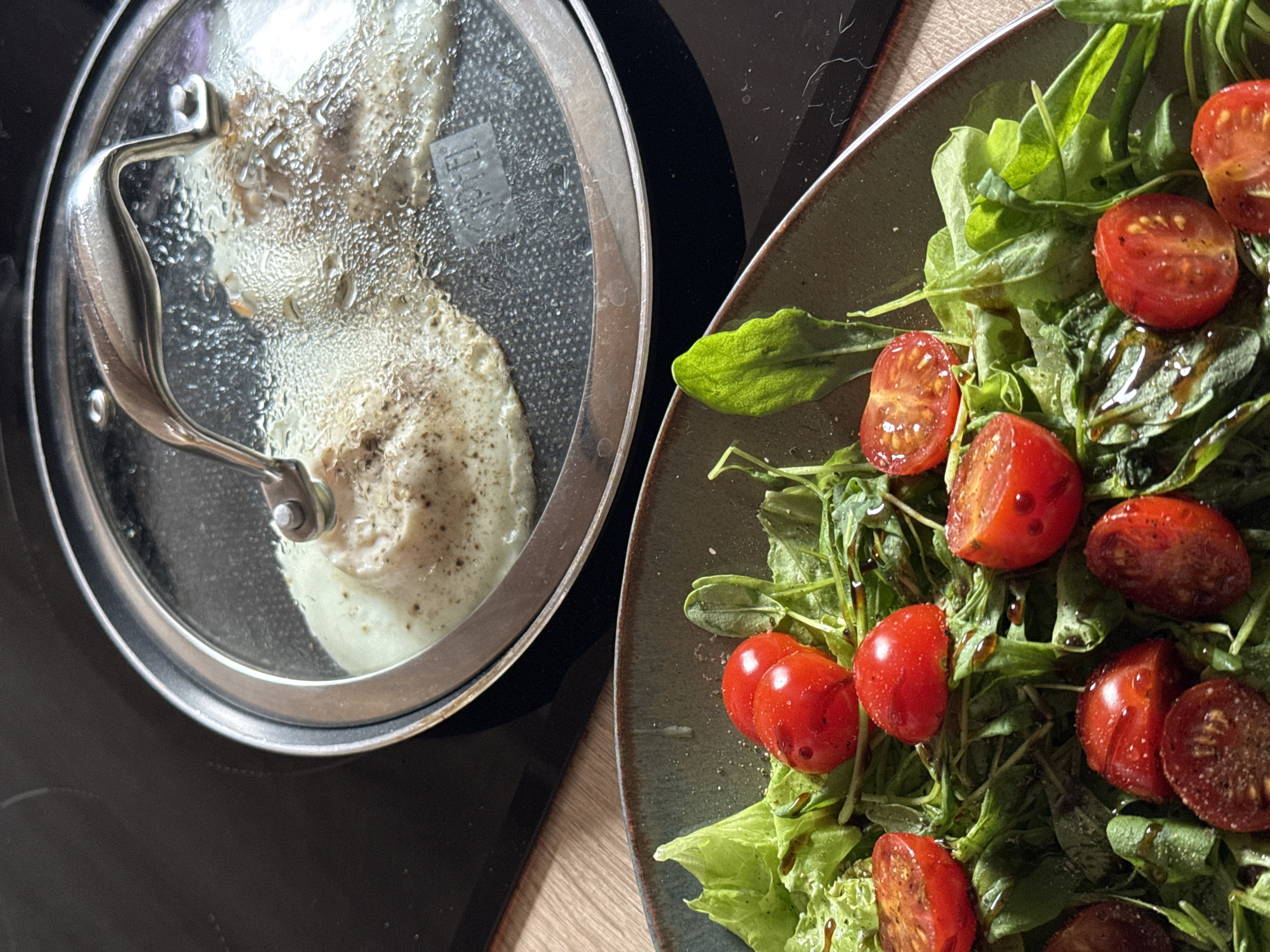 The image depicts a close-up shot of a meal being prepared. On the left, there is a frying pan with a clear glass lid, which is covered in condensation. Inside the pan, there is a fried egg seasoned with pepper. To the right of the frying pan, there is a plate of salad. The salad appears to consist of lettuce and arugula leaves, halved cherry tomatoes, and a drizzle of balsamic glaze. The frying pan is on a black cooking top and the plate is on a wooden table. There is no information to suggest that the image was taken in Krasnoyarsk, Russia.