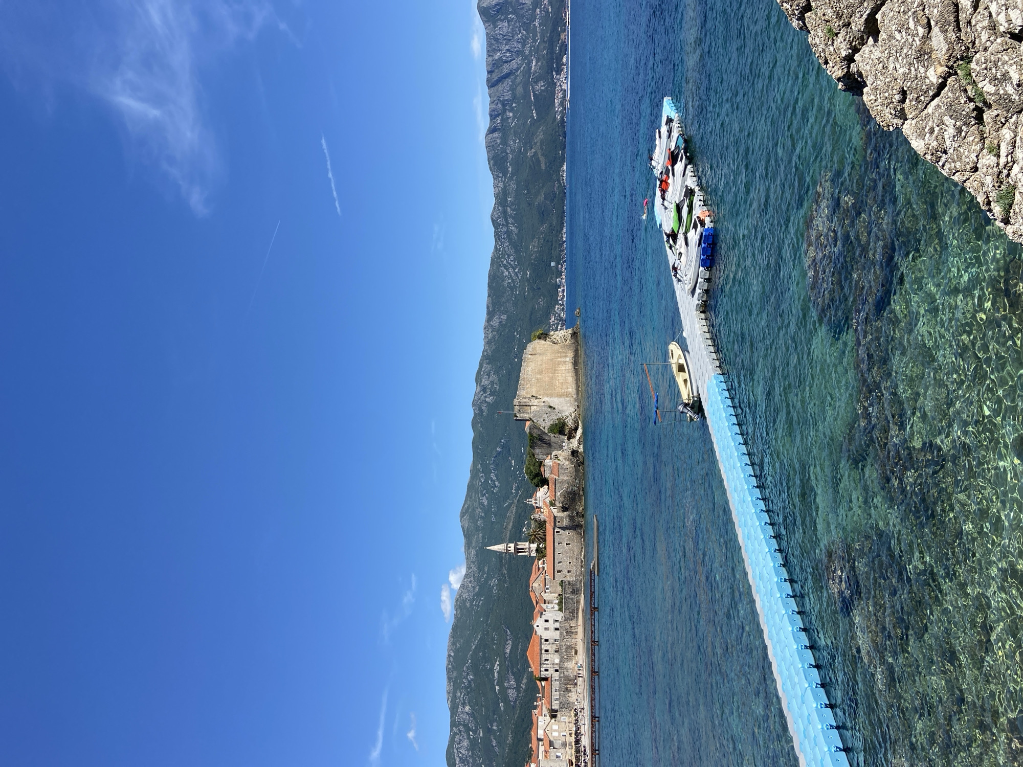 The image shows Budva, Montenegro, under a clear blue sky with scattered clouds. The camera looks down on the scene, showing a coastal landscape featuring turquoise water, a long floating dock extending from a rocky shoreline, and a town nestled against a backdrop of mountains.
The floating dock has a blue-colored base, leading to a white platform with colorful objects, possibly inflatables or watersport equipment. A small boat is docked alongside. The clear water reveals submerged rocks and marine details.
The town features buildings with orange roofs, with a church steeple rising above the structures. The mountains behind the town are covered with greenery. The scene appears serene, capturing a tourist spot in clear, sunny conditions.