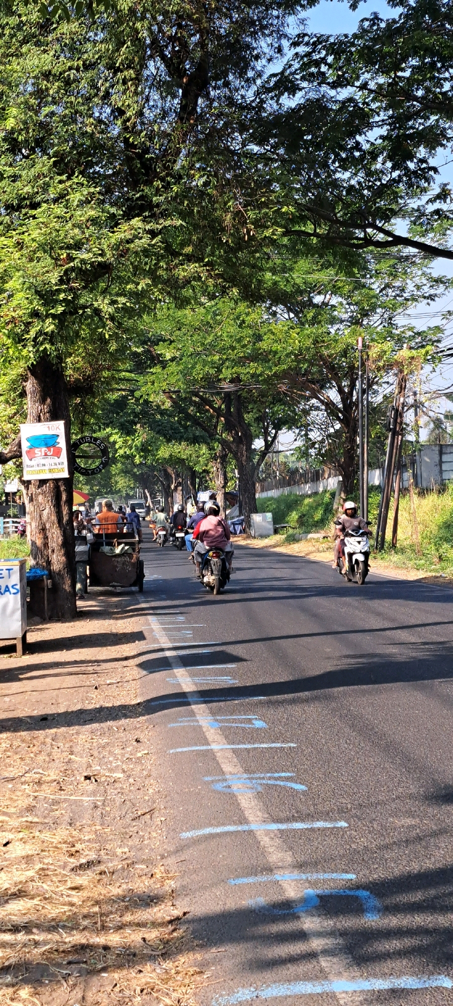 The image captures a street scene in Kebon Waris, Indonesia, bathed in bright sunlight. The road is lined with mature trees, their dense green canopies creating dappled shade on the asphalt. Several people are on motorcycles, some traveling in the same direction, while others appear to be stopped or moving at a slower pace. A man is seated in a two-wheeled cart pulled by a motorcycle.
On the left side of the road, a large tree trunk stands prominently, bearing a sign with text and a graphic. Further down the road, there are various structures and roadside stalls. The road itself has blue markings painted intermittently along its surface, suggesting a temporary or localized designation. The background shows more trees and some buildings. The sky is clear and blue.