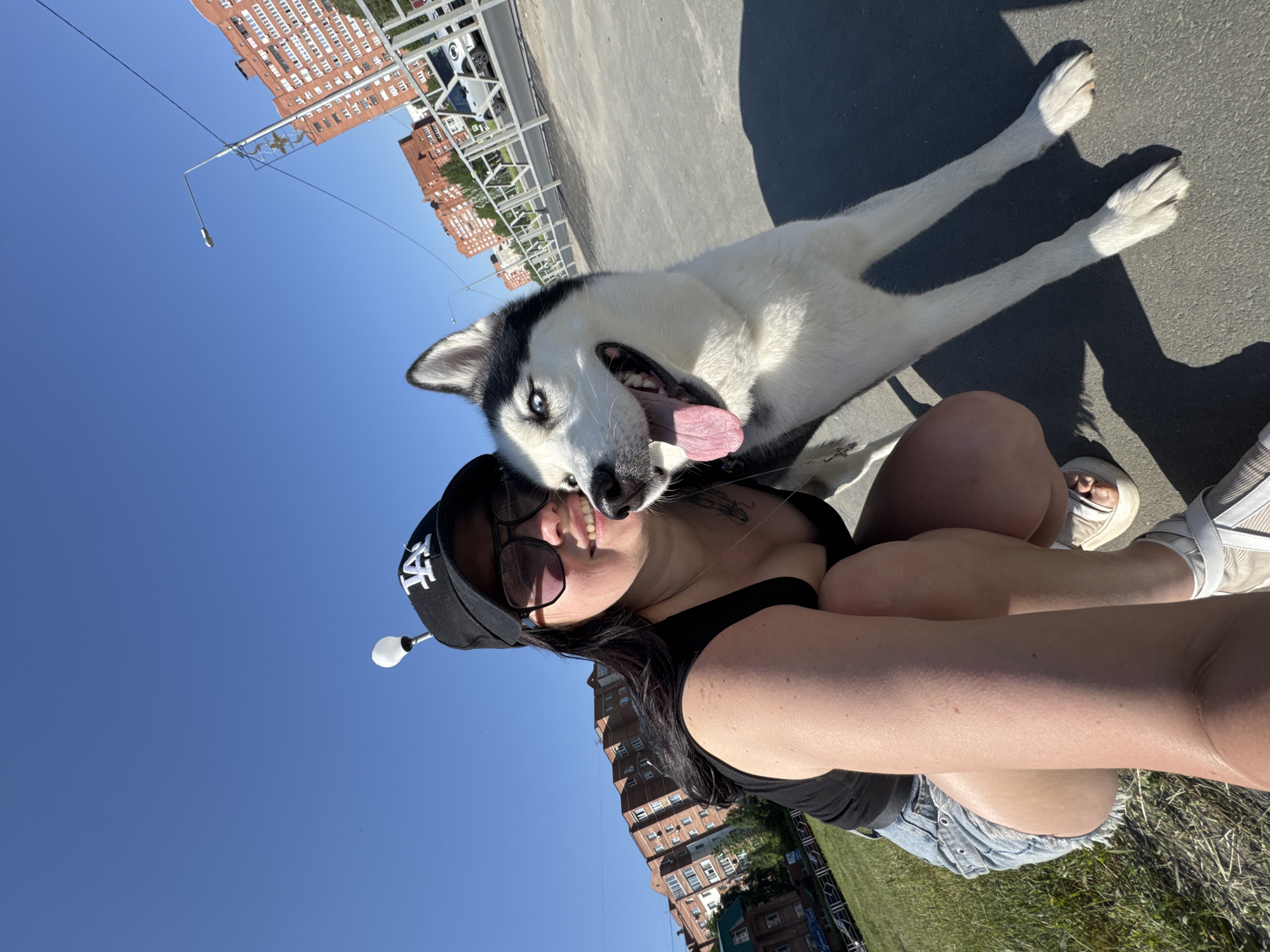 A woman and a husky-type dog are photographed outside during daylight. The woman, wearing a black tank top and jean shorts, sits on the ground while holding the camera, and the husky rests on the ground. The dog has a black and white coat with light-colored eyes. The woman is wearing sunglasses and a baseball cap. The background features urban structures including a multi-story building, an overpass, and a clear blue sky.