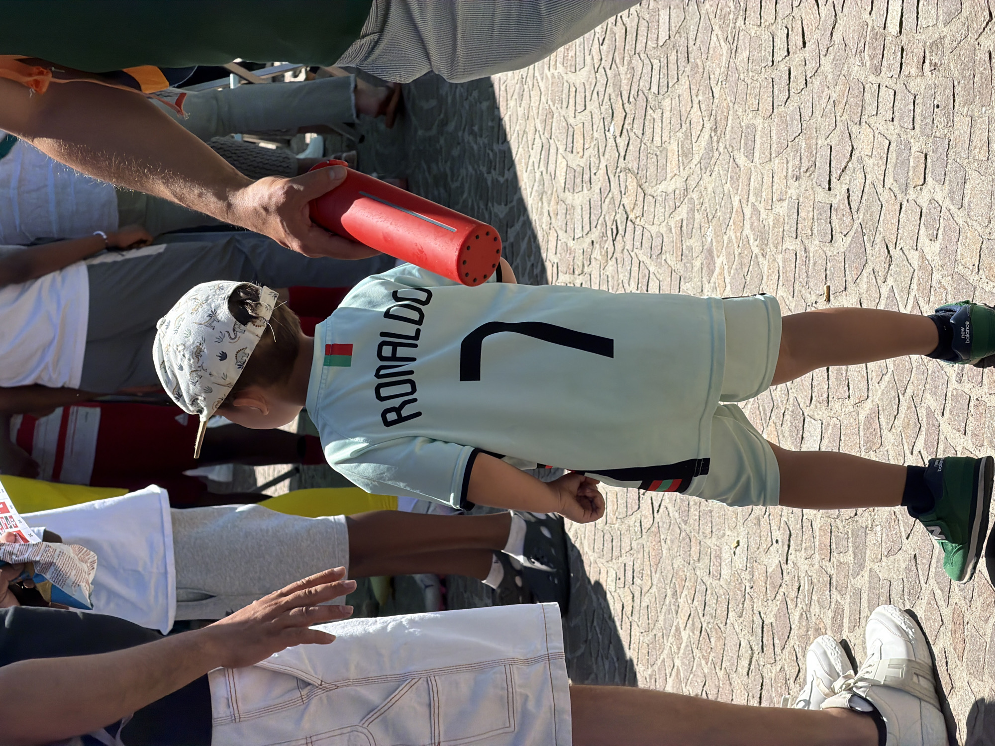 A child wearing a light blue soccer jersey with the name "RONALDO" and the number 7 on the back, along with matching shorts, is standing outdoors on a paved surface of beige and brown stones. He is also wearing a hat with a light-colored pattern. An adult's arm holds a red cylindrical object near the child. Other people are present, with parts of their clothing and bodies visible in the background. The environment appears to be a public space. There is no definitive information to identify the location as Fentange, Luxembourg, so this detail cannot be forensically confirmed from the visual evidence.
