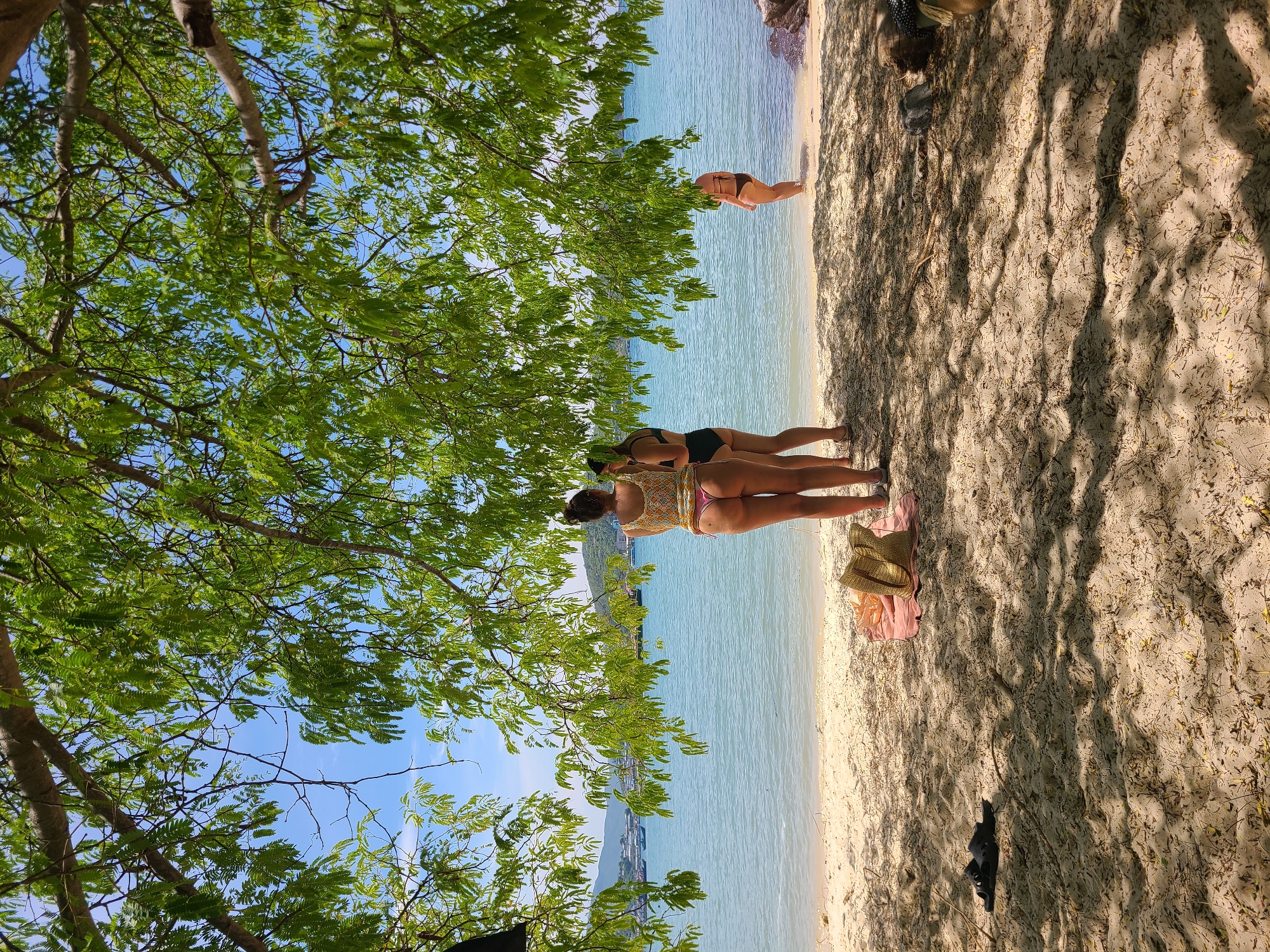 The image depicts a serene beach scene in Nha Trang, Vietnam, viewed from beneath the shade of a leafy tree. In the foreground, the sandy beach is dotted with fallen leaves and twigs, with a pair of dark sandals placed on the left. Further back, a patterned beach towel and a straw bag are visible on the sand.
Three women are present on the beach. Two are standing near the water's edge, facing away from the camera. The woman on the left wears a yellow patterned bikini top and bottoms, while the woman to her right is in a black bikini. A third woman, also in a dark bikini, is standing further in the water on the right side of the frame.
The turquoise ocean stretches out before them, meeting a clear blue sky. In the distance, the coastline is dotted with hills and buildings, and several boats are anchored in the bay. The overall atmosphere is one of relaxation and enjoyment of a tropical setting.