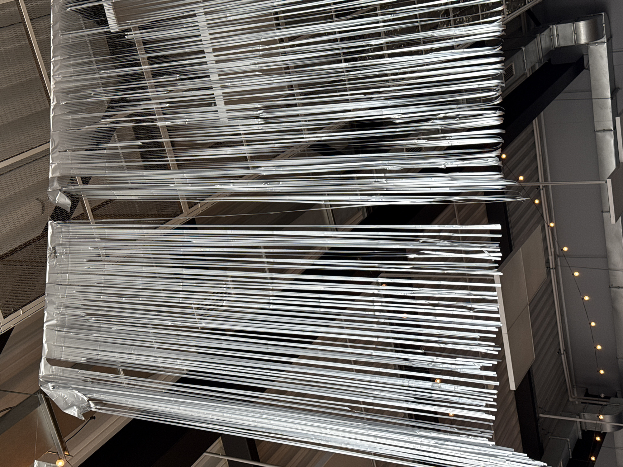 A low-angle shot of an indoor architectural setting features a hanging art installation. The installation is composed of numerous horizontal, parallel strips of a white or silver material, appearing to be suspended from the ceiling in two distinct tiers. The strips are tightly packed together, creating a dense visual pattern. The top tier of the installation is slightly elevated compared to the bottom tier, with a small gap separating the two.
The background reveals glimpses of the building's infrastructure, including a metal grid structure, ductwork, and exposed piping. A string of small, round lights is visible along a section of the ceiling. The lighting is generally dim, but reflects off the metallic strips of the art installation.
The location is not evident from the image. The location in the city of Votkinsk, Russia is not evident from the image.