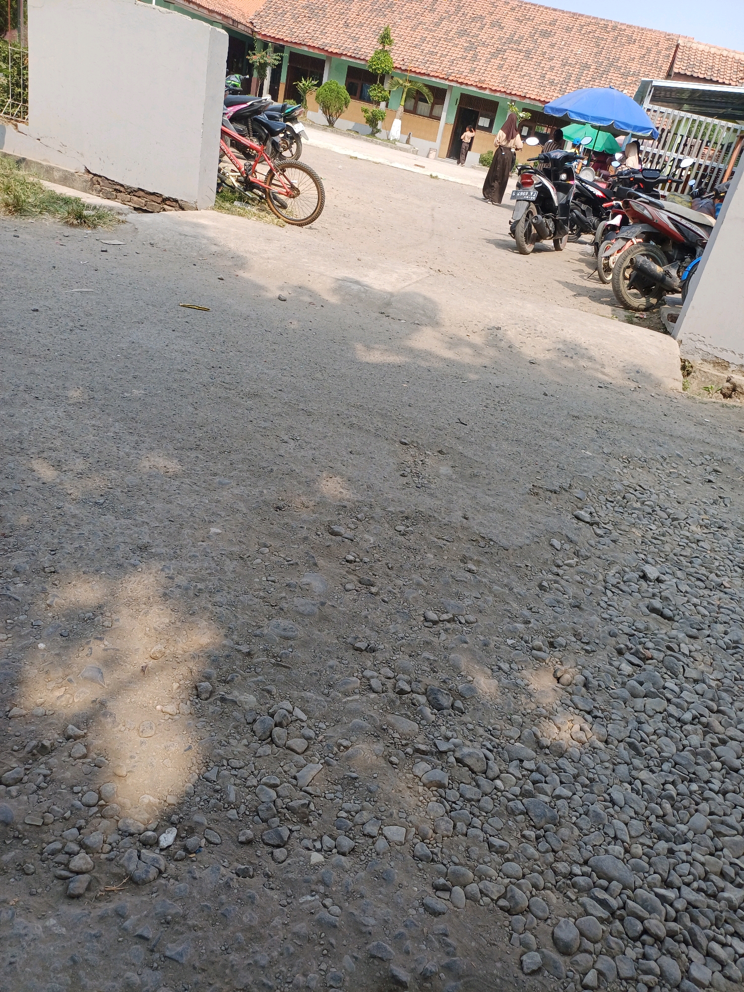 The image depicts an outdoor scene in Kersana, Indonesia, likely near a school building with a tiled roof. In the foreground, a rough, gravelly path occupies much of the frame, with shadows cast from an unseen source. To the left, a white wall partially obscures a red bicycle. Further back, a row of parked motorcycles is visible, some covered by blue and green umbrellas. Several individuals are present, including two women in dark clothing and headscarves standing near the motorcycles. One woman appears to be looking towards the building entrance, where another person in a light-colored shirt and dark pants stands. The environment appears sunny, with the sky visible in the upper right portion of the image.