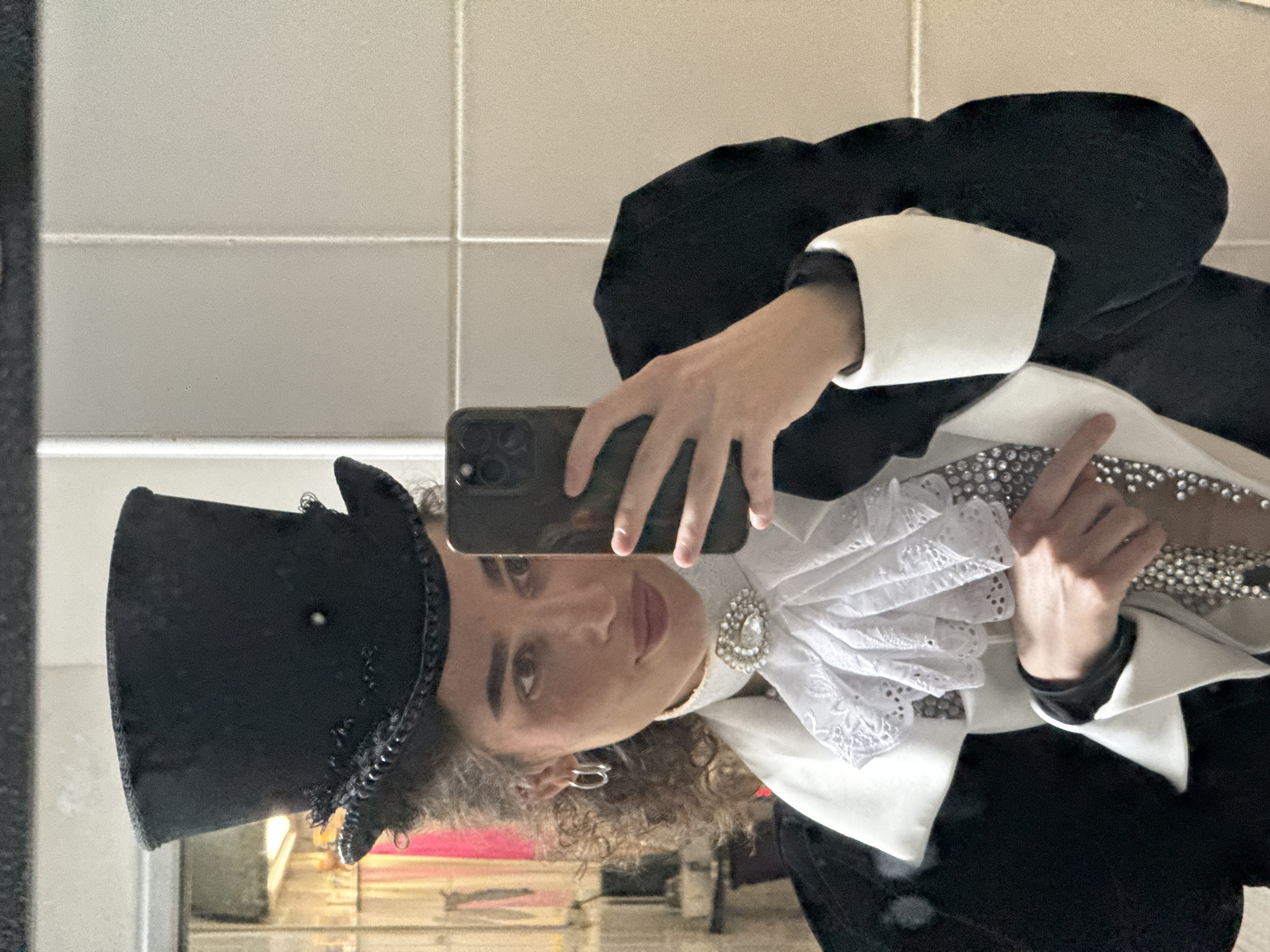 A person is taking a selfie in a mirror, wearing a black top hat and a white ruffled ascot with a jeweled brooch. The person is also wearing a black jacket with white cuffs and trim, and a beaded necklace. The background appears to be a tiled bathroom. There is no indication of location in Kazan, Russia.