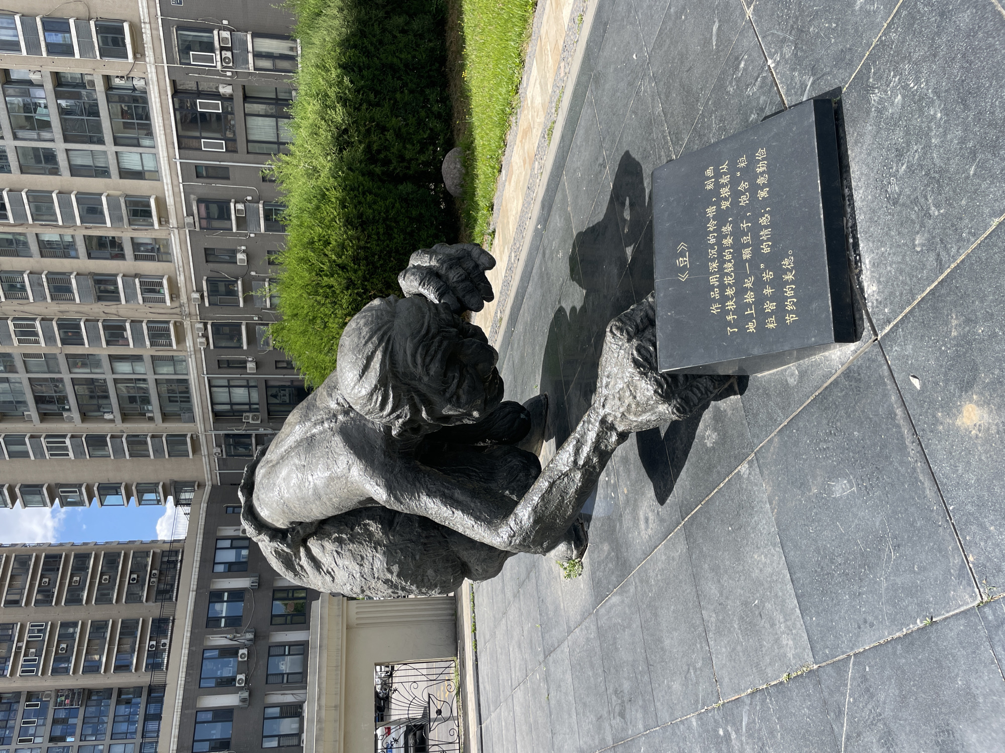 Here's a detailed description of the image:
The image shows a bronze statue of an elderly woman crouching, likely picking something up from the ground. She is wearing what appears to be traditional clothing, and her features are heavily wrinkled, suggesting age. The statue is positioned on a paved area made of dark gray tiles. Next to the statue is a rectangular black plaque with writing in Chinese characters. The text on the plaque is an inscription describing the statue as a depiction of an elderly woman picking up a bean, meant to evoke feelings of reverence for labor and the virtues of thrift and frugality.
Behind the statue and plaque, there's a neatly trimmed green bush. Further in the background, a large, multi-story building with numerous windows is visible, suggesting an urban or residential area. The sky is partially visible through the upper parts of the building, indicating a clear day.
The image was taken in the Changping District of China, as indicated by the plaque. The scene appears to be in a park or public space.