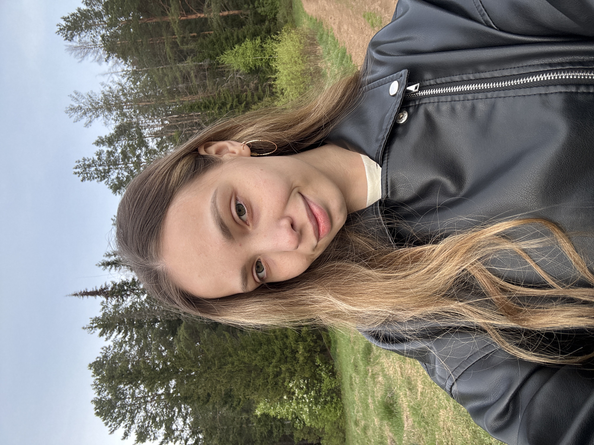 A young woman with long, light brown hair is posing outdoors. She is wearing a black leather jacket and a hoop earring in her left ear. The background consists of trees and grass. The location is Прикамский, Russia.