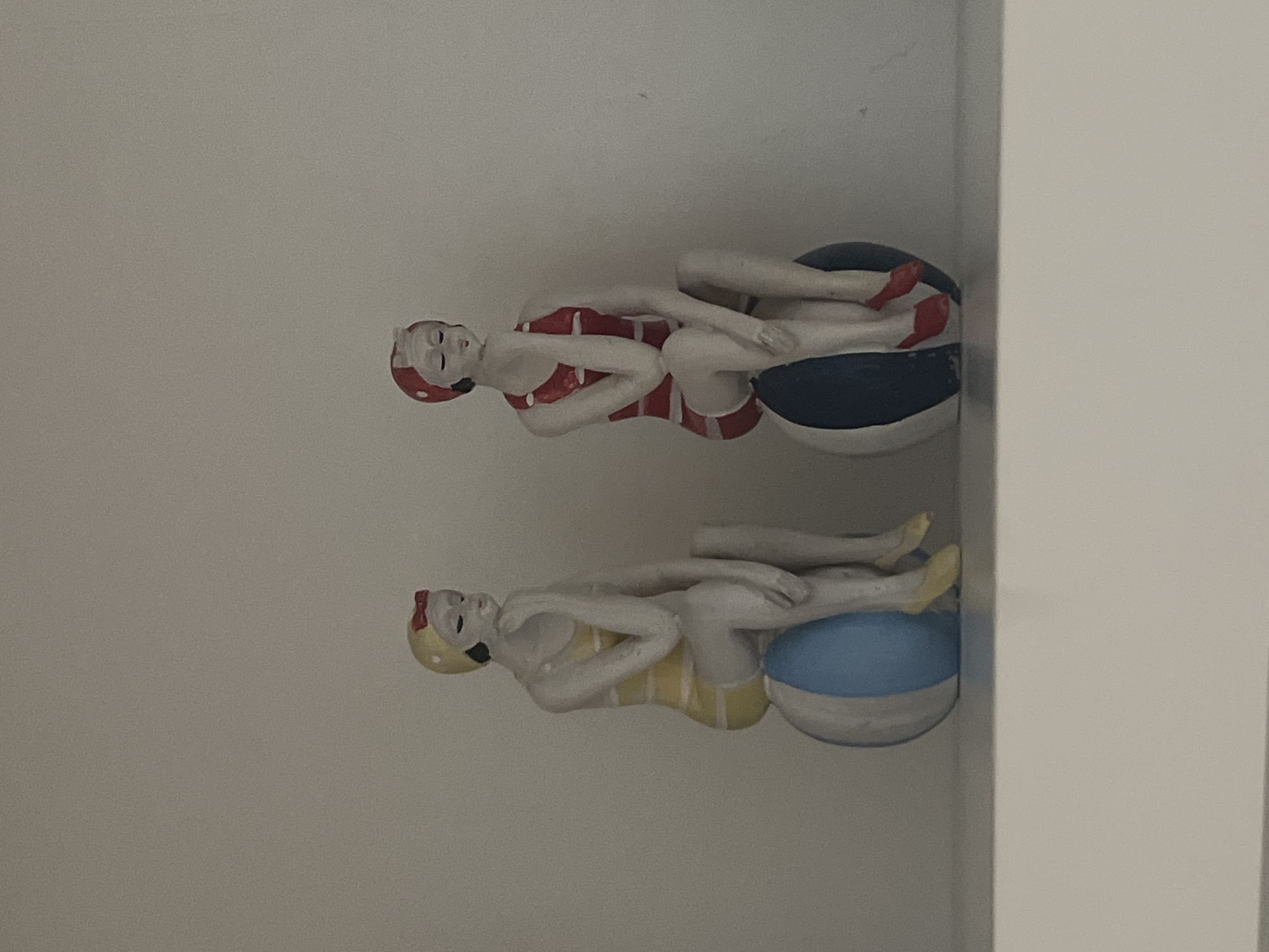 Two decorative figurines of women in vintage swimsuits are displayed on a white shelf against a plain background. The figurine on the left wears a yellow and white striped swimsuit and cap, sitting on a blue and white striped ball. The figurine on the right wears a red and white striped swimsuit and cap, also sitting on a blue and white striped ball. Both figurines are depicted with closed eyes and their hands resting on their chins, suggesting a contemplative pose. The scene appears to be indoors, likely in a home or shop. There is no indication of the location being in the city of Ko Samui District, Thailand.