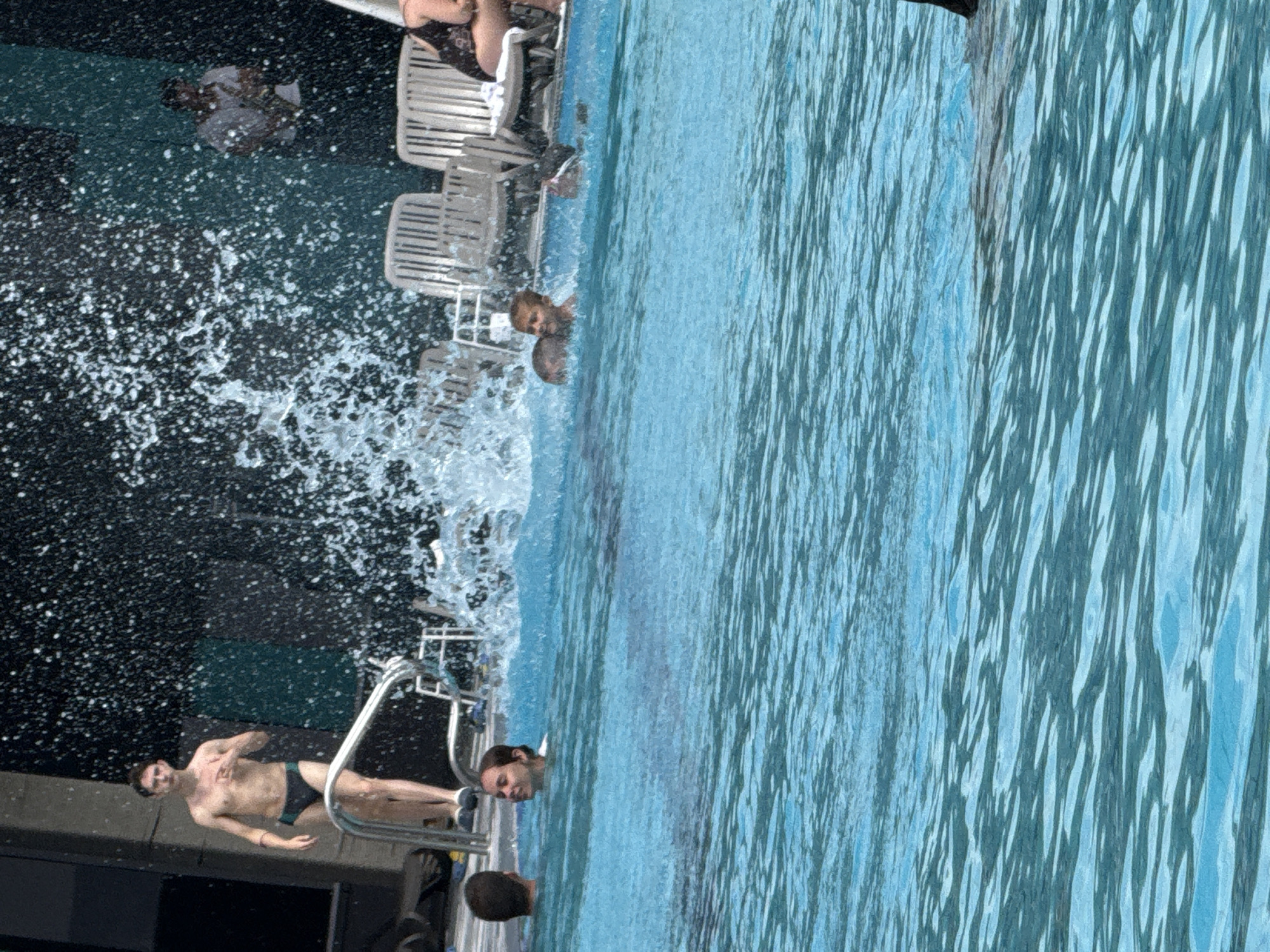 The image depicts a lively scene at an outdoor swimming pool, likely located in Yalta, Ukraine, given the context. In the foreground, the bright blue water of the pool is rippled, suggesting activity. Several people are visible enjoying the water. On the left, a young man stands on a diving platform, wearing black swim trunks and pointing towards the water. He appears to be in the process of diving or has just finished. Further into the pool, a young woman with dark hair is swimming, looking towards the right of the frame. To her right, a young boy, also with light brown hair, is in the water, looking ahead with a slight smile. There are hints of other individuals in the water, some submerged or partially visible. In the background, white lounge chairs are arranged on a deck surrounding the pool. Beyond the pool area, a dark, textured wall creates a backdrop, with a man in a white shirt and dark pants visible in the far background, seemingly engaged with something on the wall. A significant amount of water spray is present in the mid-ground, indicating dynamic splashing, possibly from diving or play. The overall atmosphere is one of recreation and enjoyment on a sunny day.