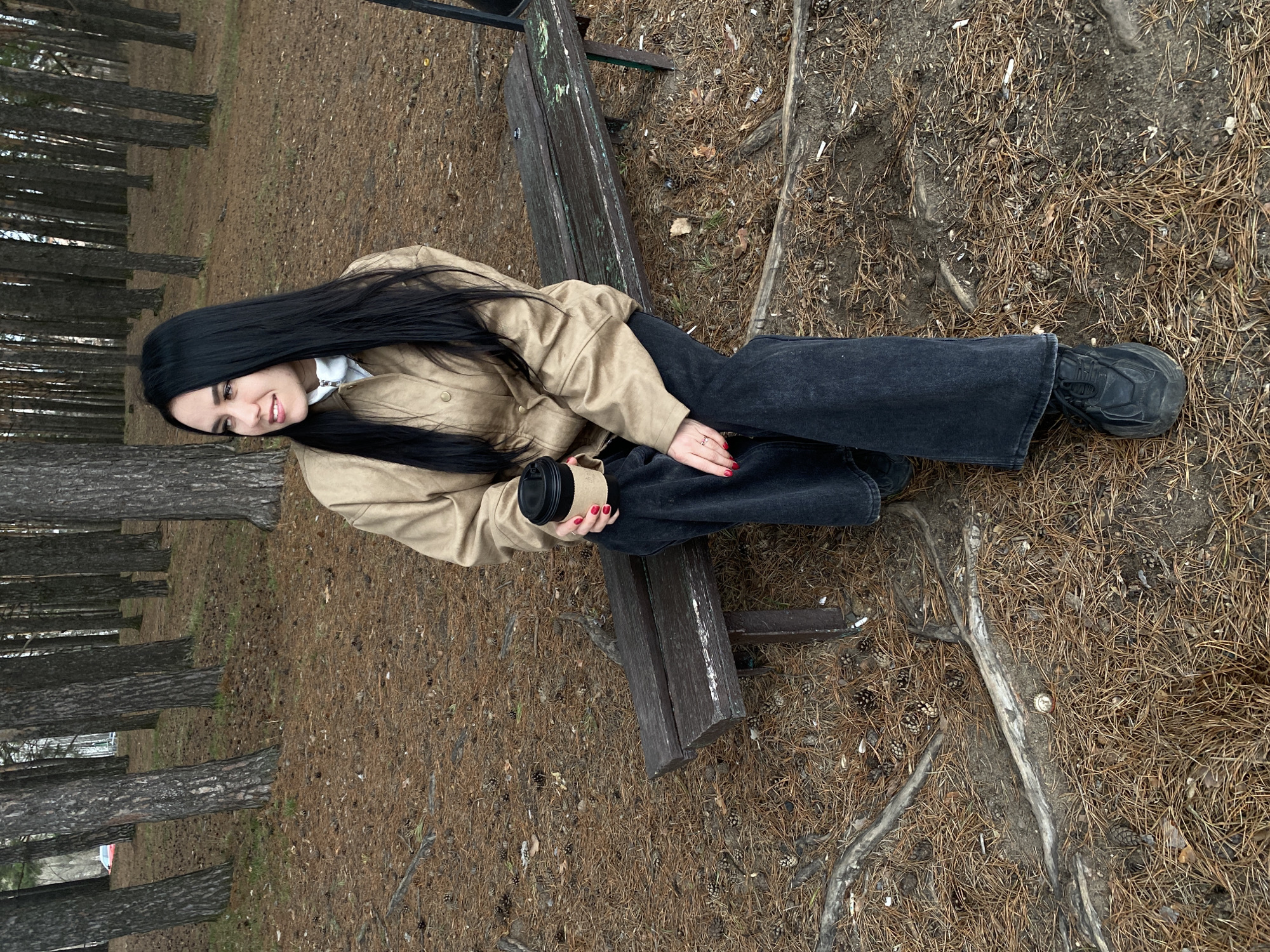 A young woman is seated on a weathered wooden bench in a pine forest or park. She has long, straight black hair and is wearing a light tan or beige jacket over a white top, dark gray or black wide-leg jeans, and black sneakers. Her fingernails are painted red, and a silver ring is visible on her right hand. She is holding a dark-colored disposable coffee cup with a light brown paper sleeve in her left hand, which rests on her lap. The woman is smiling gently and looking directly towards the camera.
The ground around the bench and extending into the background is covered extensively with pine needles, pine cones, and exposed tree roots. Tall, slender pine trees line the background, creating a dense wooded environment. The lighting appears soft and diffuse, suggesting an overcast day. No specific time of day is indicated beyond daylight. The setting aligns with the provided context of a forest in Chaikovsky, Russia. No discernible text is visible on the coffee cup or elsewhere in the image.