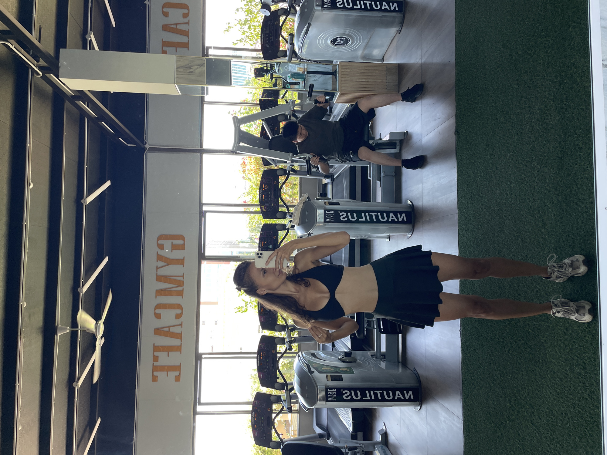 A woman stands in a gym, taking a mirror selfie. She is wearing a black sports bra and a black skirt. Behind her, treadmills and weight machines are visible. A man is exercising on a weight machine in the background. The gym appears to be located in Ham Thang Ward, Vietnam, as evidenced by the "GYMCAFE" signage visible on the wall. The environment is well-lit with natural light streaming in from large windows.