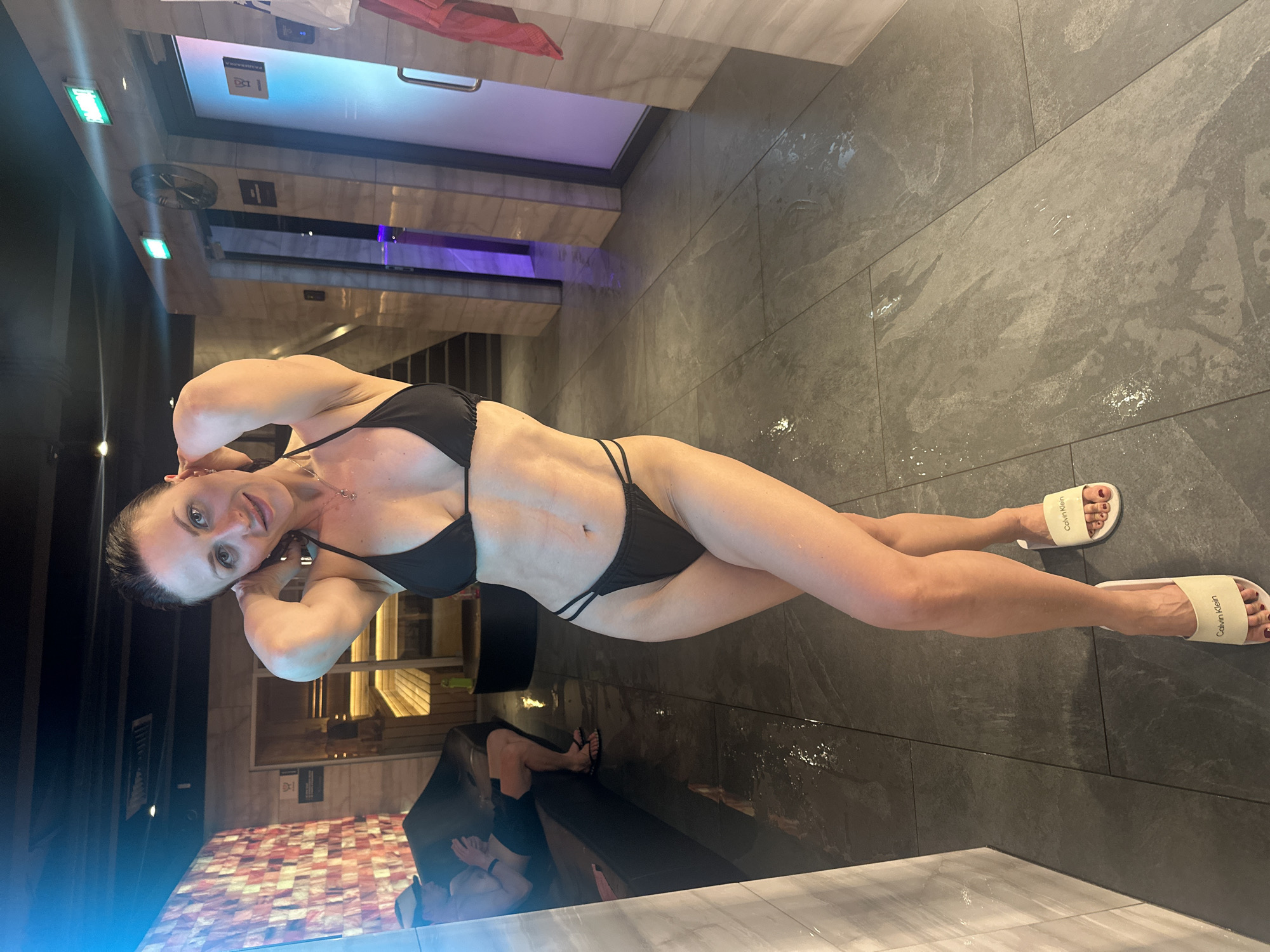 A woman stands in a modern bathroom, likely a spa or wellness center, wearing a black bikini and white Calvin Klein branded slide sandals. The setting features gray tile flooring and walls. The lighting appears bright and artificial, possibly from recessed lights or illuminated panels. A sauna is visible in the background, characterized by its wooden interior and a color-changing light strip along the top of the entryway. Another individual is reflected in the water. The location is most likely in Saint Petersburg, Russia.