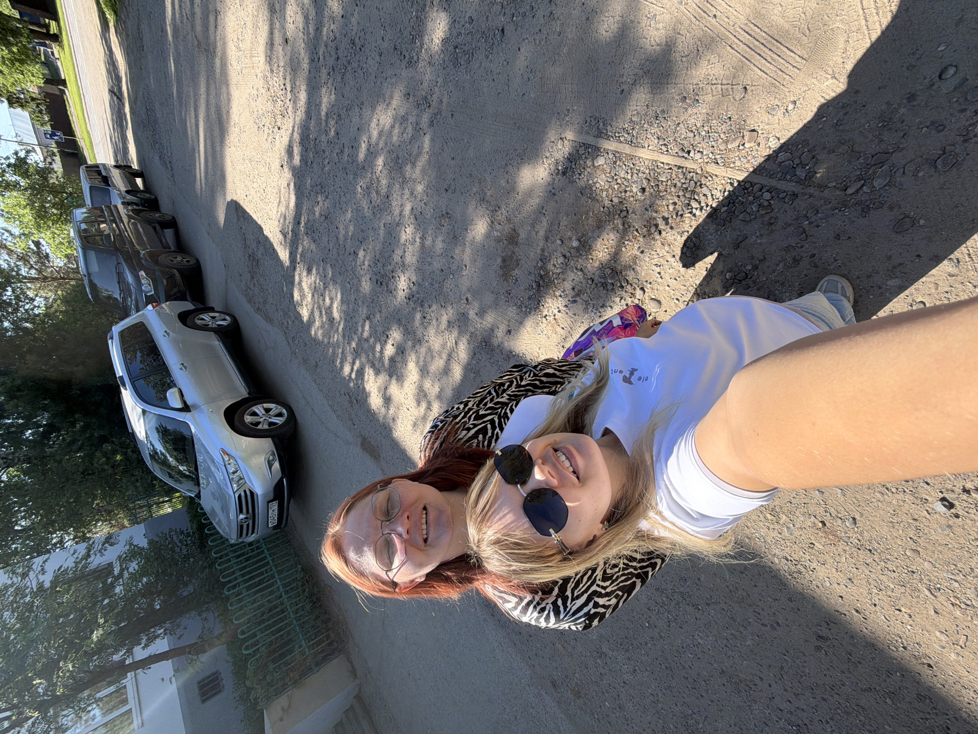 Two women are posing for a selfie outdoors. One woman has long, red hair and is wearing glasses, while the other has long, blonde hair and is wearing sunglasses. They are both smiling. They are wearing what appears to be a zebra-striped shawl. A parking area with parked cars, and greenery is visible in the background. The location is Chaikovsky, Russia.