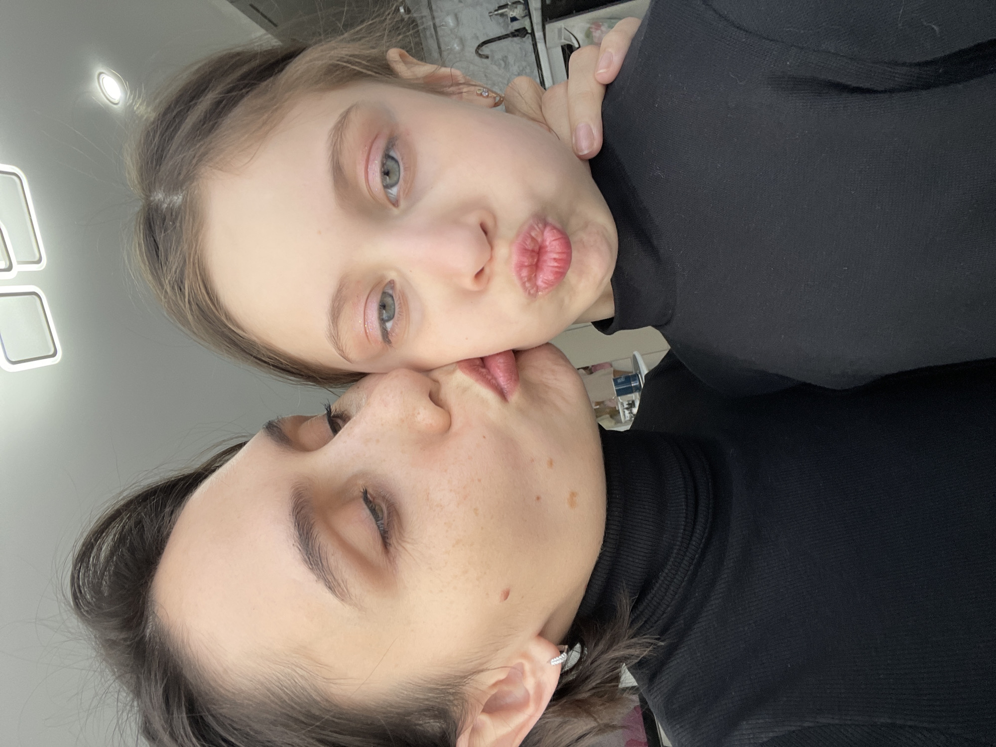 A close-up shot captures two individuals, likely a mother and daughter, in an affectionate pose. The daughter, positioned above, puckers her lips towards the mother's face. Both are wearing black turtleneck sweaters. The mother's eyes are closed. The background includes a light fixture and indistinct objects, suggesting an indoor setting. There is no information within the provided image that would indicate this photo was taken in Izhevsk, Russia.