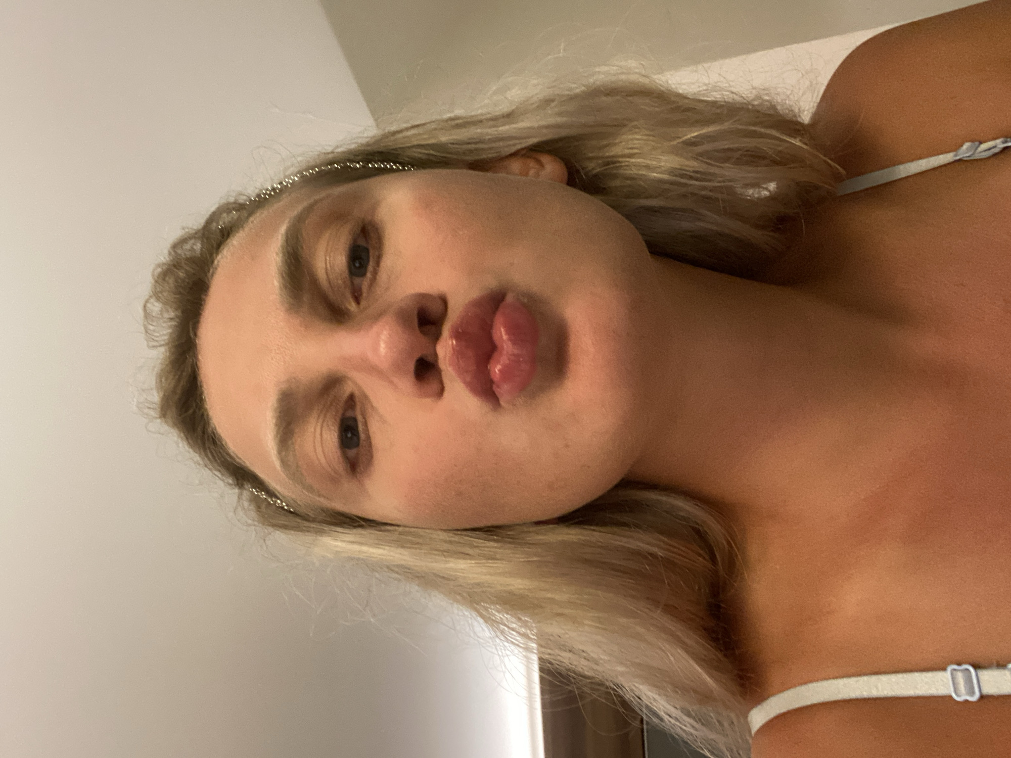 A young woman with blonde hair is captured from the chest up, looking directly at the camera with her lips puckered. She is wearing a light-colored strap, possibly from a camisole or bra. A delicate, beaded headband adorns her hair, partially obscured by her blonde locks. The background is a plain, light-colored wall, suggesting an indoor setting. No specific location in Aksu, Turkey, can be identified from the visual information present in the image.