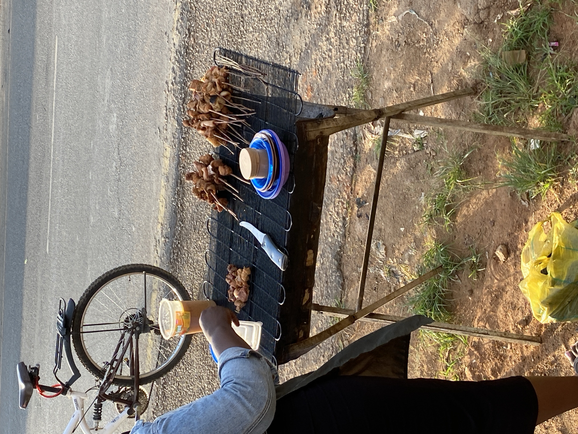 A top-down shot shows a street food vendor next to a road, likely in Bukuru, Nigeria. The vendor's metal stand displays grilled meat skewers, stacked blue and purple plates, and a knife. A plastic container is held above the stand. A bicycle leans against the stand on the left. The ground around the stand is dirt with patches of grass. A yellow plastic bag sits on the ground on the right. The road is paved. The sky appears clear, indicating sunny conditions. A person wearing a denim jacket and black skirt is visible.