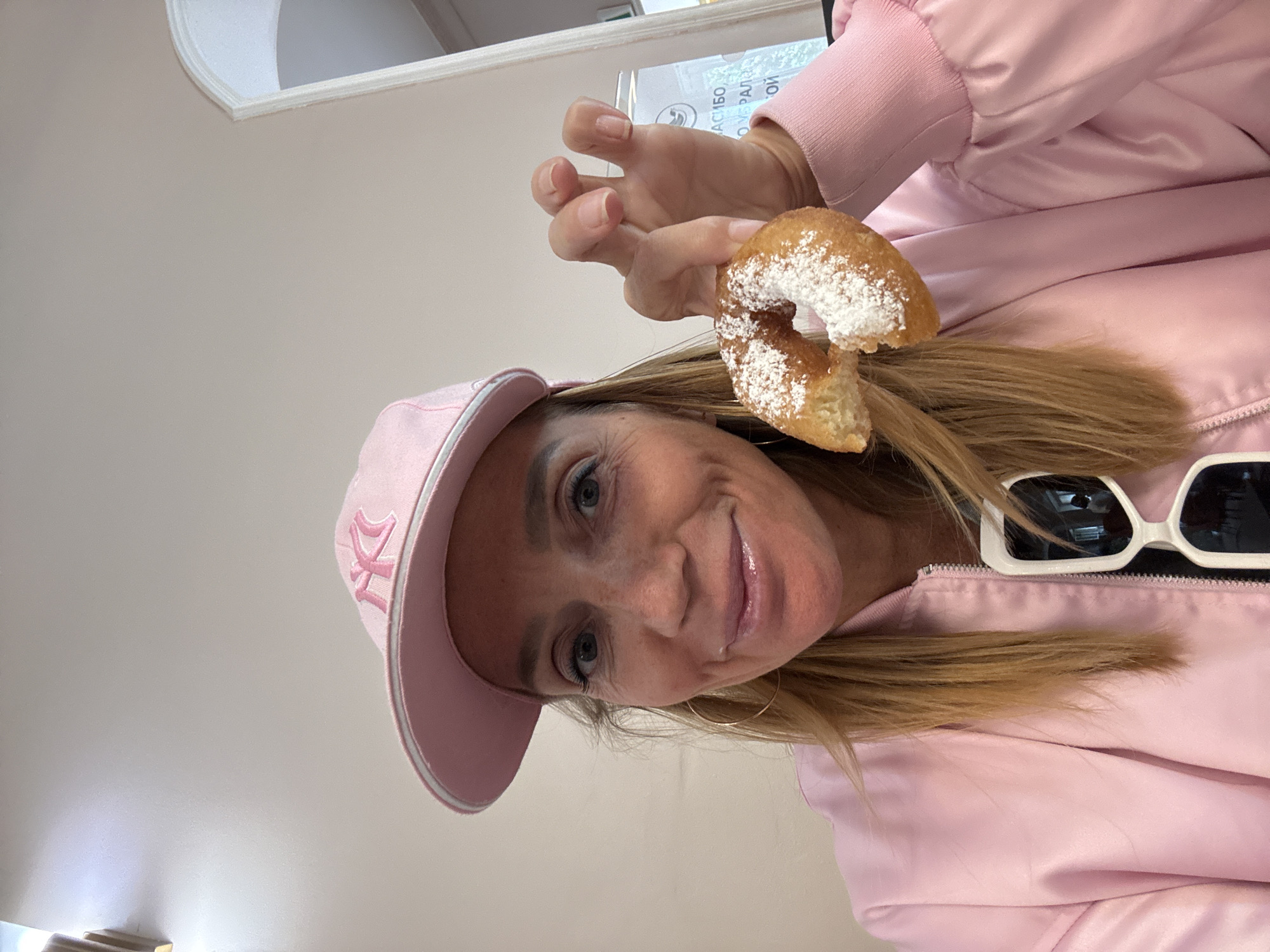 A woman is captured in a close-up, indoor shot, smiling directly at the camera while holding a half-eaten, powdered sugar-dusted donut. She has light-colored eyes and long, light brown or blonde hair. She is wearing a light pink baseball cap with a white embroidered logo, possibly "DK," and a shiny, light pink bomber jacket. White-framed sunglasses are resting on her jacket collar. Large hoop earrings are visible.
The background is a plain, light beige or off-white wall. In the upper left, a subtle architectural detail with a curved recess can be seen. Behind her hand, a white sign with partially visible Cyrillic text is present. Recognizable parts of the text include "АСИБО" (likely part of "Спасибо," meaning "thank you") and "О УРАЛ" (possibly referring to the Ural region or a company). The woman's expression is cheerful and engaging, suggesting a pleasant moment. The setting appears casual, and the lighting indicates daytime. This is consistent with an indoor setting in Saint Petersburg, Russia.