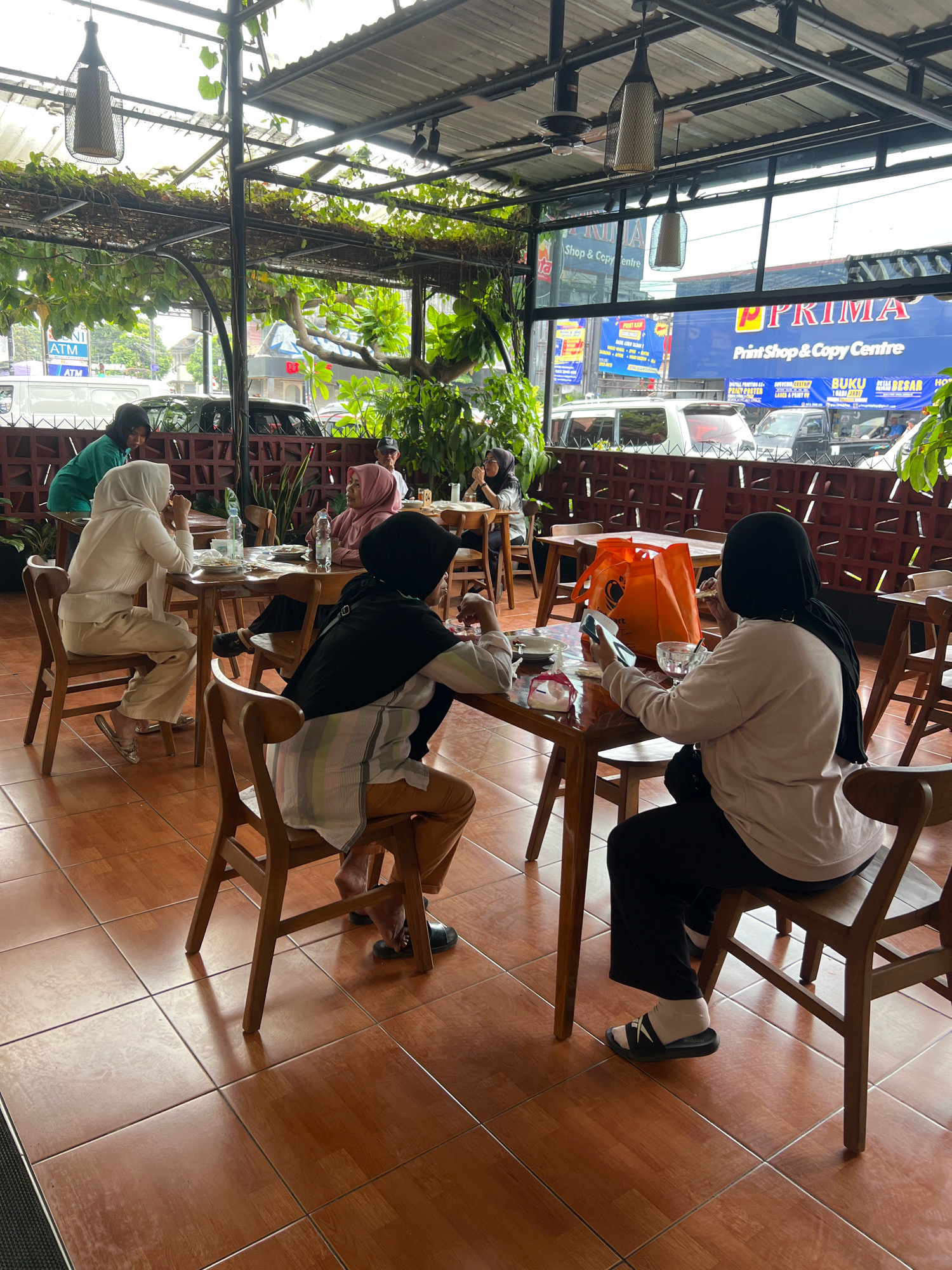 The image depicts an outdoor dining area of a cafe or restaurant, possibly located in Yogyakarta, Indonesia. Several people, primarily women wearing hijabs, are seated at wooden tables, engaged in eating and conversation. The environment features a covered seating area with a rustic, natural aesthetic, including hanging plants and wooden beams. In the background, there are signs of urban activity, with vehicles and buildings visible, suggesting a location within a city. The floor is tiled with a warm, reddish-brown color. One woman in the foreground is looking at her phone, while others are interacting with each other or their food. An orange shopping bag is placed on the table. The overall scene suggests a casual dining experience.