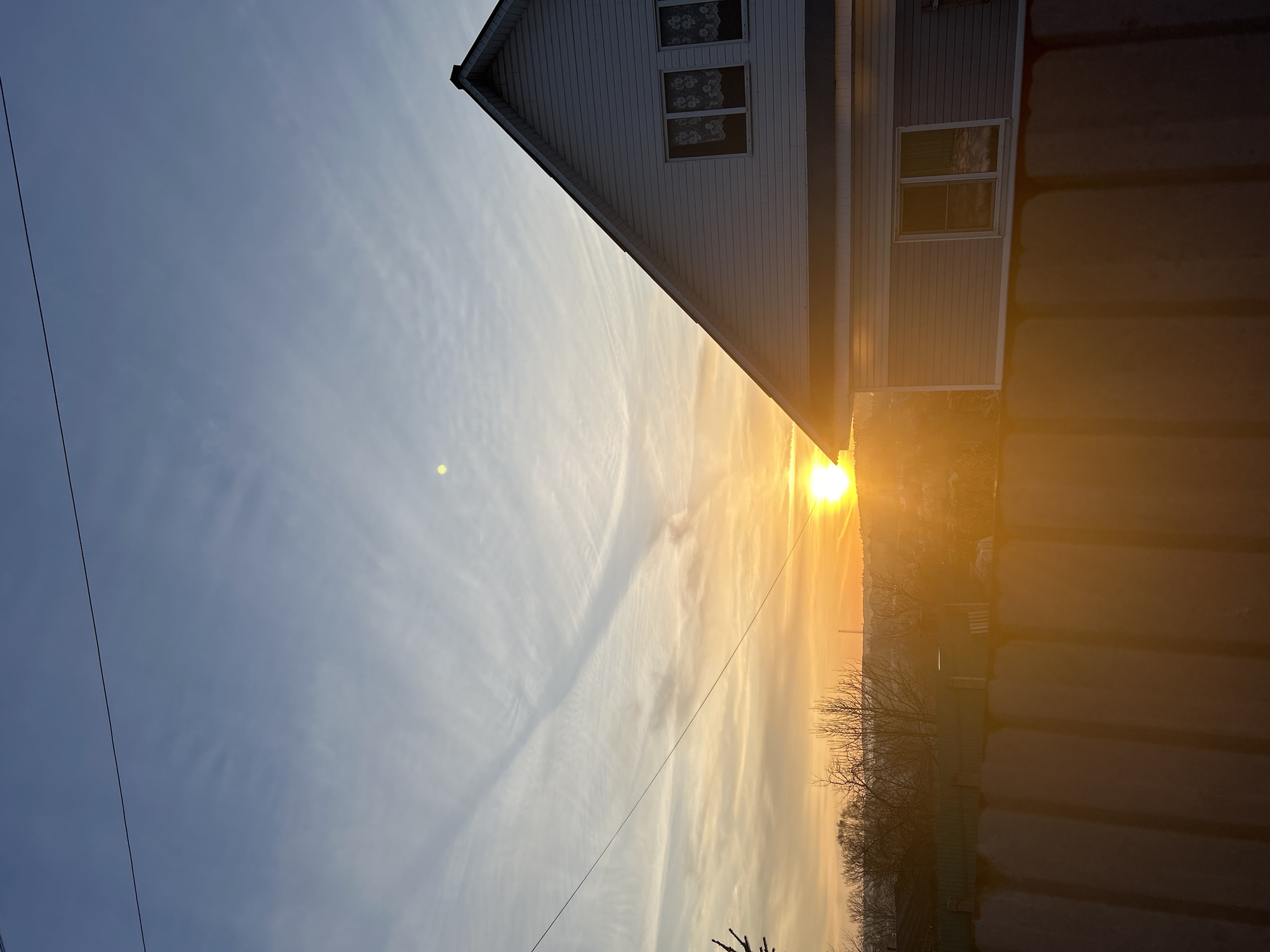 The image shows a low-angle shot of a house with a white facade and several windows. The sun is setting or rising, creating a bright glare near the horizon. A power line is visible in the sky. Bare trees are silhouetted against the bright sky. A solid, dark-colored structure or barrier runs along the bottom right edge of the frame. The location is likely near Фоки, Russia.