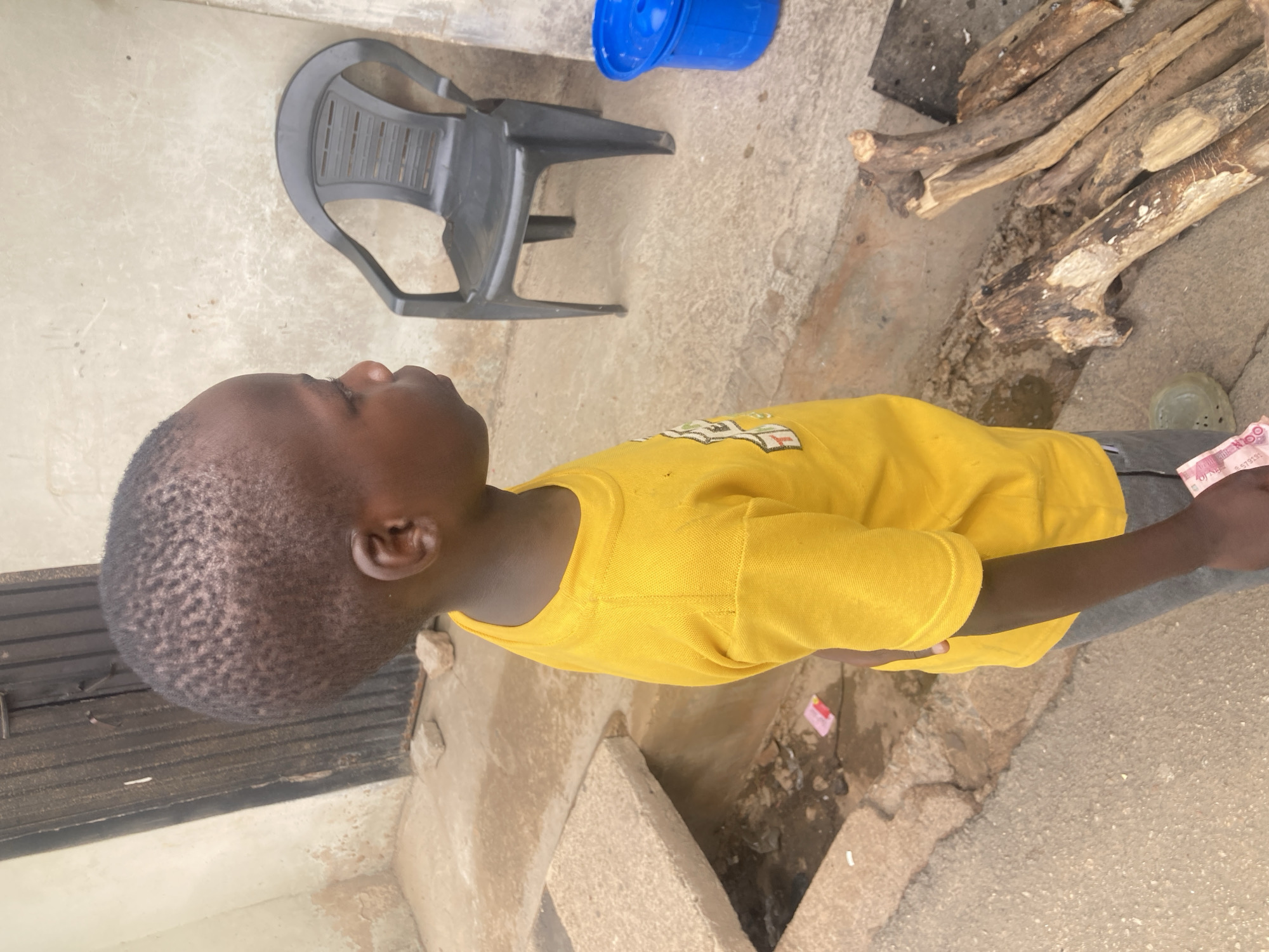 A young Black child wearing a yellow shirt stands outdoors on a concrete surface. The child appears to be holding a stack of banknotes in one hand. To the child's left is a concrete step or curb. In the background are a black plastic chair, a blue plastic bucket, and a stack of firewood. The location is possibly within the city of Jos, Nigeria, but this is only suggested.