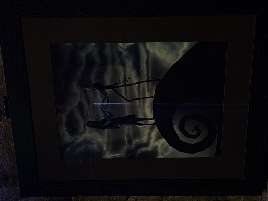 A framed image depicts a scene from "The Nightmare Before Christmas." Jack Skellington, a skeletal figure, stands on a swirling hilltop next to Sally, a ragdoll girl. The background is a cloudy, dreamlike sky. The frame has multiple layers, a light interior and dark outer. The photo is somewhat dark and has a light glare across the middle.