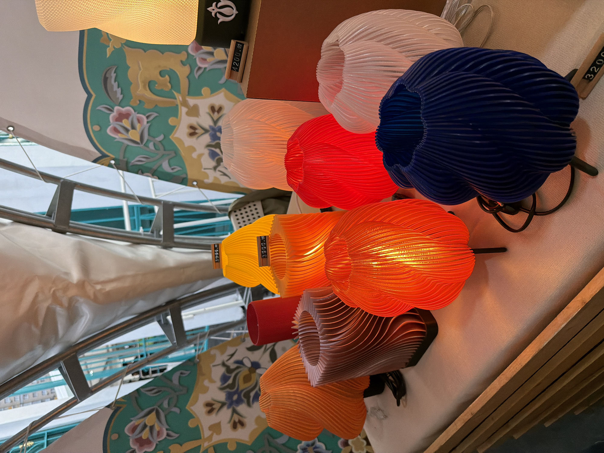 An indoor shot displays a collection of colorful, petal-shaped lamps arranged on a white surface, possibly a table or shelf. The lamps are made of intricately folded material, creating a textured and layered effect. From left to right, there are lamps in orange, a reddish-brown swirl pattern, yellow, red, white, and a deep blue. Behind the lamps, a large, tent-like structure with a white, translucent fabric is visible, supported by a metal framework. Decorative panels with floral patterns in teal, yellow, pink, and blue are also part of the backdrop. A wooden price tag displaying "3900.00" is placed on the yellow lamp, and another price tag with "4200.00" is seen near a white lamp. Another wooden price tag with "3200" is visible on the bottom right. A red cylindrical object stands behind some of the lamps. The lighting is warm, highlighting the glow from within the illuminated lamps. The scene appears to be a retail display or an exhibition booth.