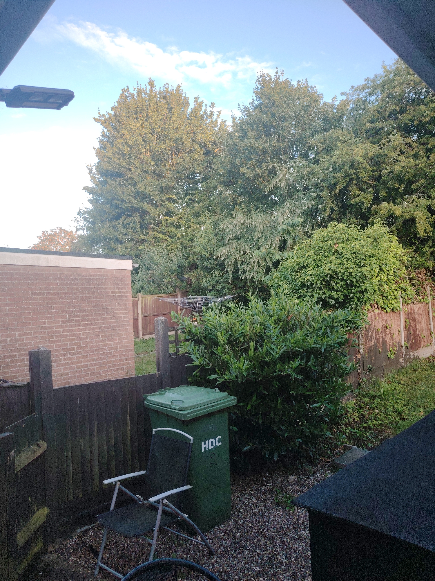 The image captures a residential backyard scene, possibly in the city of Huntingdonshire, United Kingdom, given the presence of an "HDC" labeled bin, likely referring to Huntingdonshire District Council. In the foreground, a green refuse bin with the "HDC" logo is positioned on a gravel surface. Adjacent to it is a dark-colored folding chair. A wooden fence encloses the yard, with a brick building visible on the left. Beyond the fence, a dense area of trees and foliage dominates the background, with a clear sky featuring scattered clouds overhead. A modern outdoor light fixture is mounted on the left side of the image, angled towards the sky. The overall impression is that of a functional, somewhat overgrown, outdoor space.