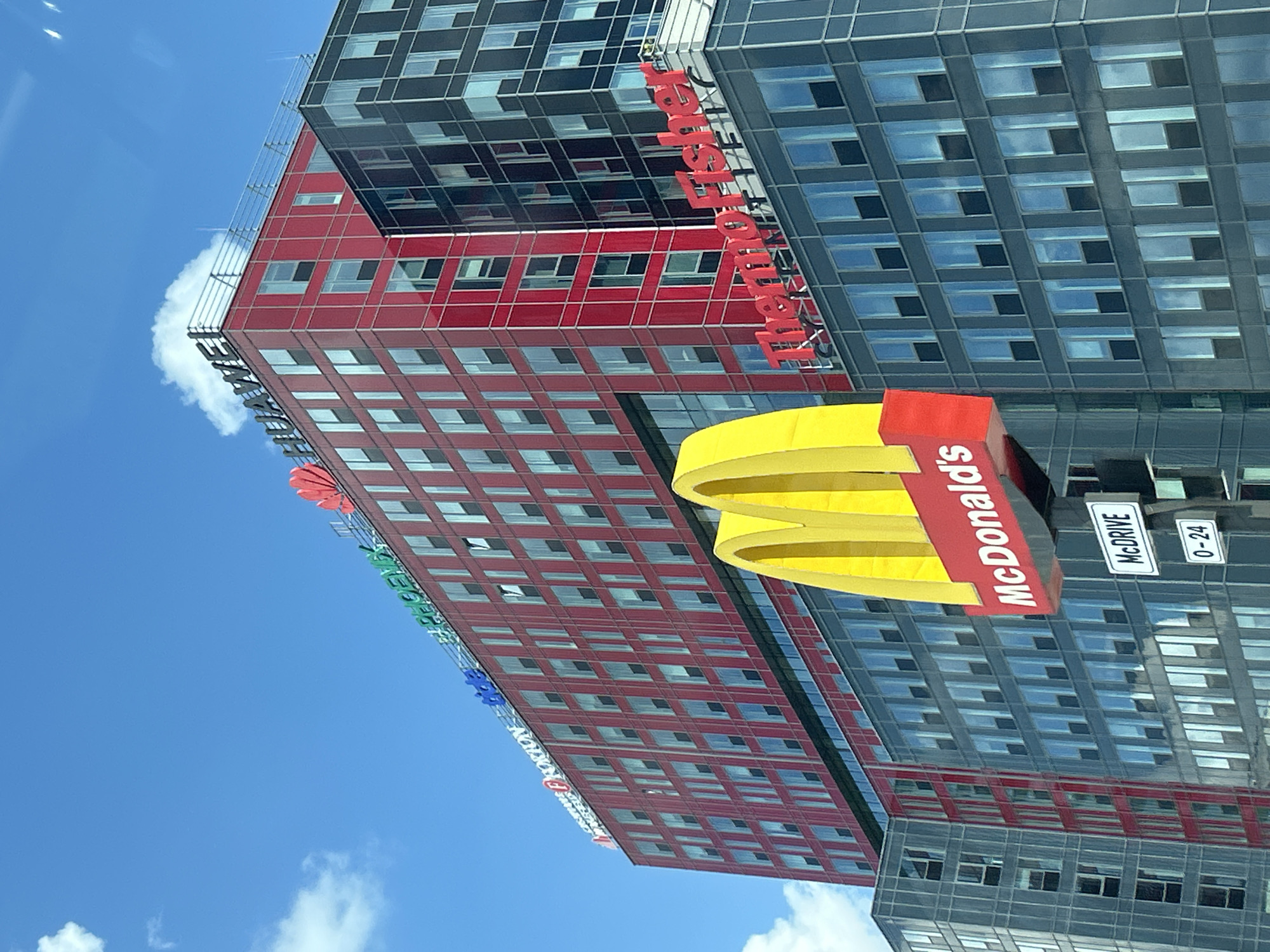 The image displays a modern urban scene featuring multiple multi-story office buildings and commercial signage under a clear blue sky with scattered white clouds. In the foreground, a prominent yellow McDonald's "M" arch logo with "McDonald's" written in white on a red background is positioned. Below this, a white sign indicates "McDRIVE" and "0 - 24" hours of operation.
Behind the McDonald's sign, a building with a blue-gray glass facade is visible. To its right and extending upwards, a large red building dominates the midground. This red building features numerous rectangular windows and displays several company logos along its upper edge, including "NORTON" (white text on a red background), "PROXION" (red text), "tide" (blue text), "PHOENIX" (green text), and "HUAWEI" (white text) with its distinctive red petal-shaped logo.
To the right of the red building, a darker, multi-story building with a grid-like facade is present, featuring numerous dark-tinted windows. On the upper section of a lower part of this dark building, the red text logo "ThermoFisher Scientific" is clearly visible. No individuals are discernible in the scene.
This location is identified as part of the Office Park Sofia complex in Mladost 4, Sofia, Bulgaria, with the specific buildings and their displayed corporate tenants corresponding to this business park.