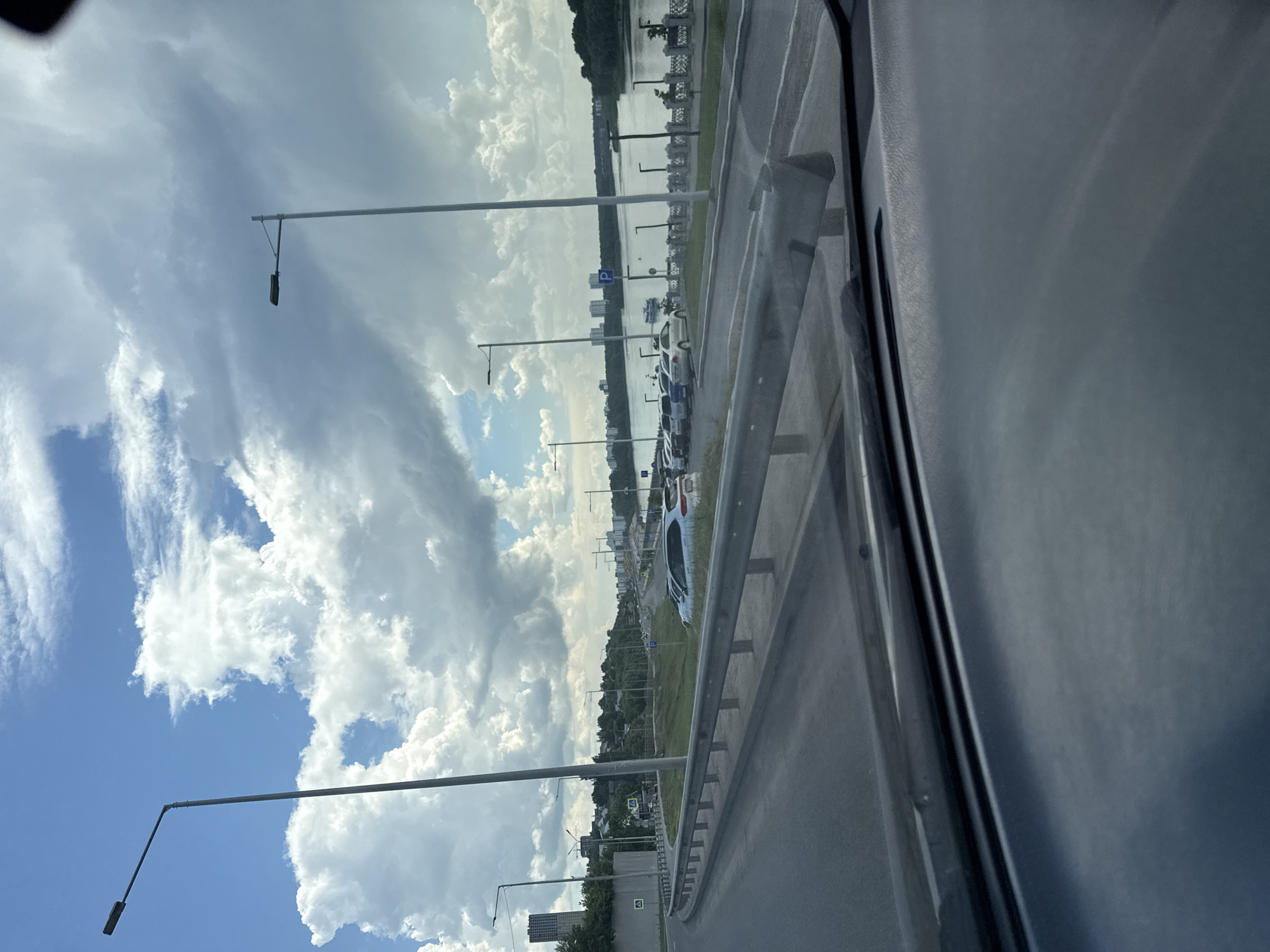 The image depicts a scene from inside a vehicle looking out at a city embankment with a river on the right. The sky is partly cloudy with large, white cumulus clouds against a blue sky. The embankment features a road, a guardrail, and a grassy slope. Several streetlights are visible along the road.
On the embankment, a line of parked cars is visible, with a white SUV in the foreground. Beyond the cars, a river flows, with a promenade or walkway along its bank. Several buildings, including tall residential structures, can be seen on the far side of the river, interspersed with trees. The overall impression is of a sunny day with dramatic cloud formations, a well-developed urban area adjacent to a body of water. The foreground of the image shows the dashboard and windshield of the vehicle from which the photo was taken.