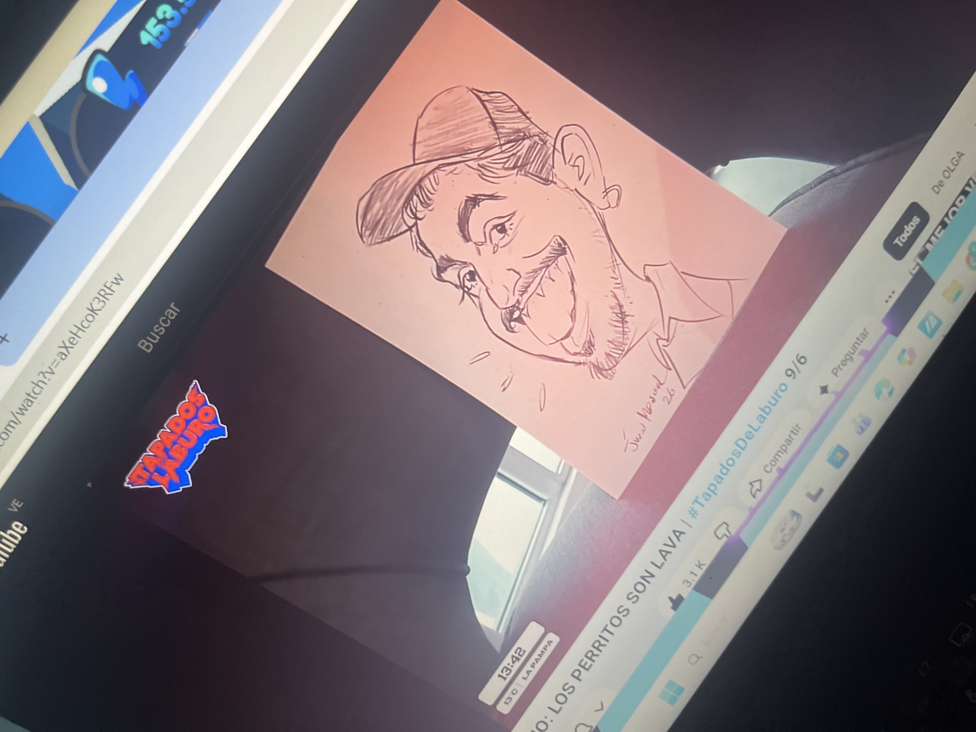 The image shows a close-up of a laptop screen displaying a YouTube video. A pink piece of paper with a caricature drawing of a person wearing a baseball cap is placed on the laptop screen, obscuring part of the video. The drawing is in black ink and features exaggerated facial features. The YouTube video title is visible as "0: LOS PERRITOS SON LAVA | #TapadosDelaburo 9/6". Below this, the current temperature is shown as 13°C in La Pampa, and the time is 13:42. The search bar displays "Buscar" and a URL fragment "v=aXehcoK3rfw". A logo that reads "TAPADOS DELABURO" in blue and red is also visible on the screen. The background of the image is dark, likely the surrounding environment of the laptop. There is no discernible information about the city of Santa Ana, Venezuela, in this image.